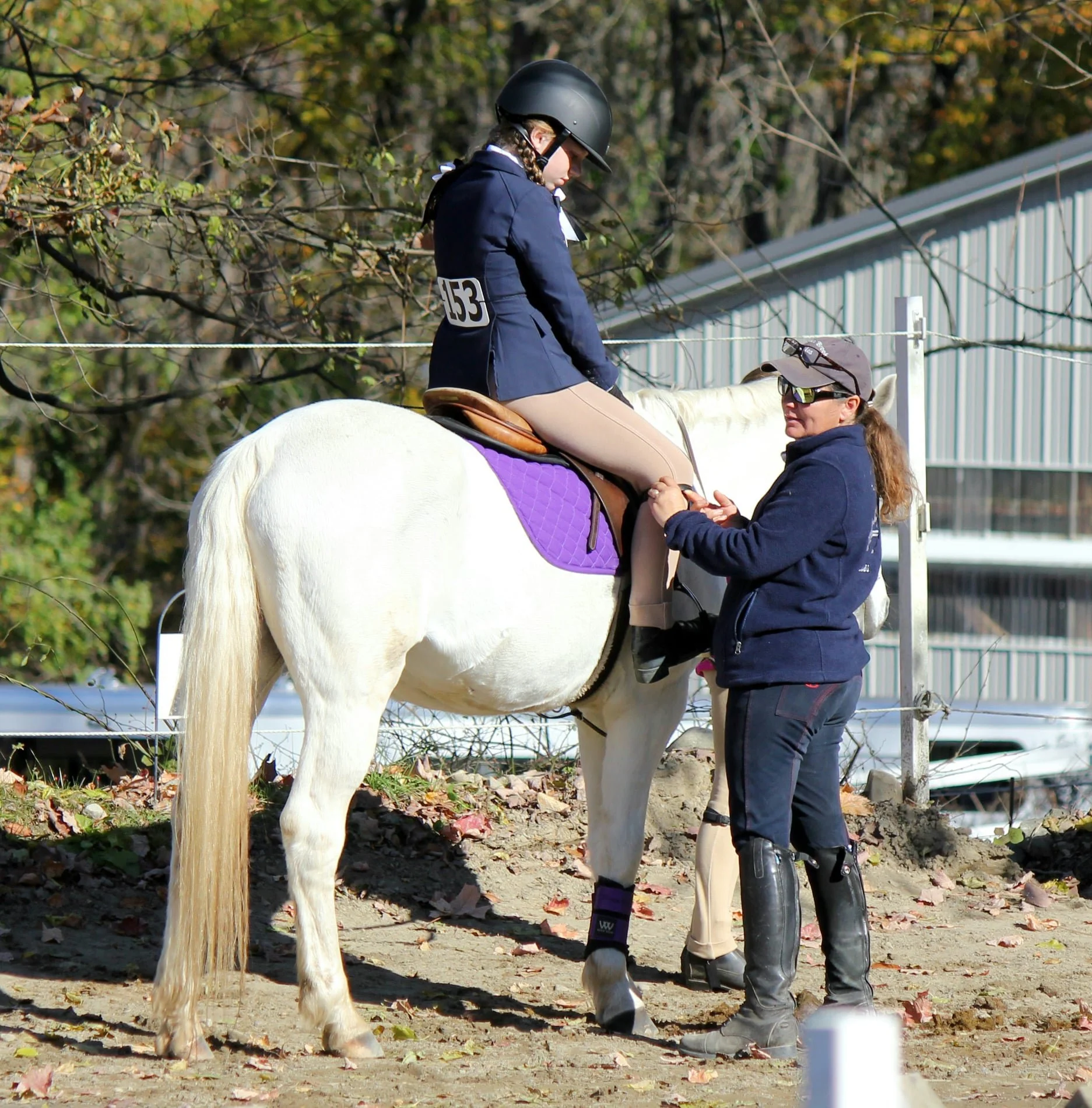 A person sits mounted on a grey horse, wearing a helmet and riding gear, with another person standing next to them helping secure the saddle. The scene takes place outdoors in a wooded area.