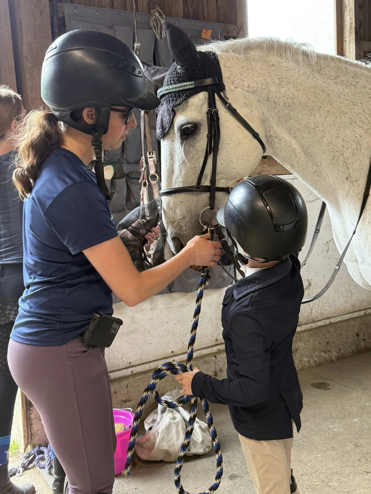 A woman and a boy wearing helmets are standing near a grey horse inside a barn. The woman appears to be helping the boy adjust the horse's harness or bridle.