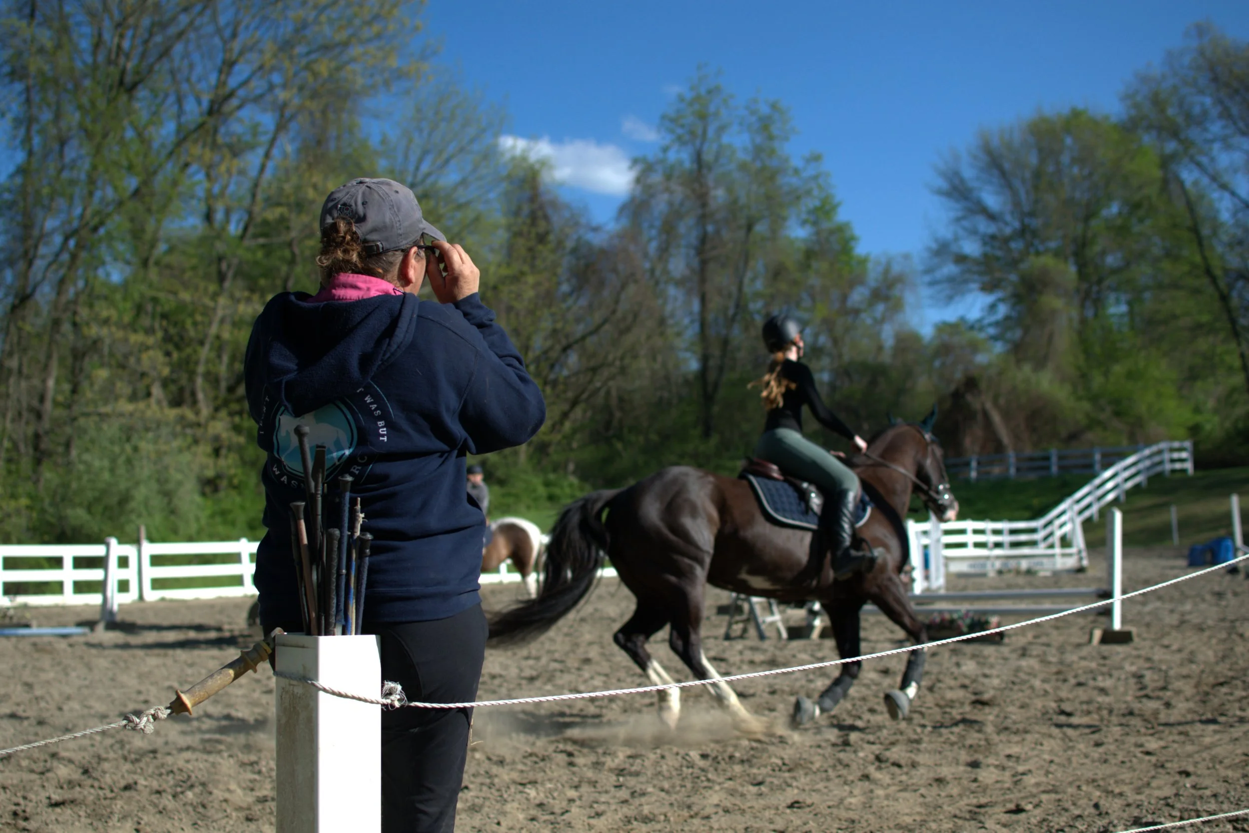 A woman with glasses, a gray hat, and a pink scarf taking a photograph or video in an equestrian riding arena. In the background, a woman riding a horse, surrounded by green trees on a sunny day.