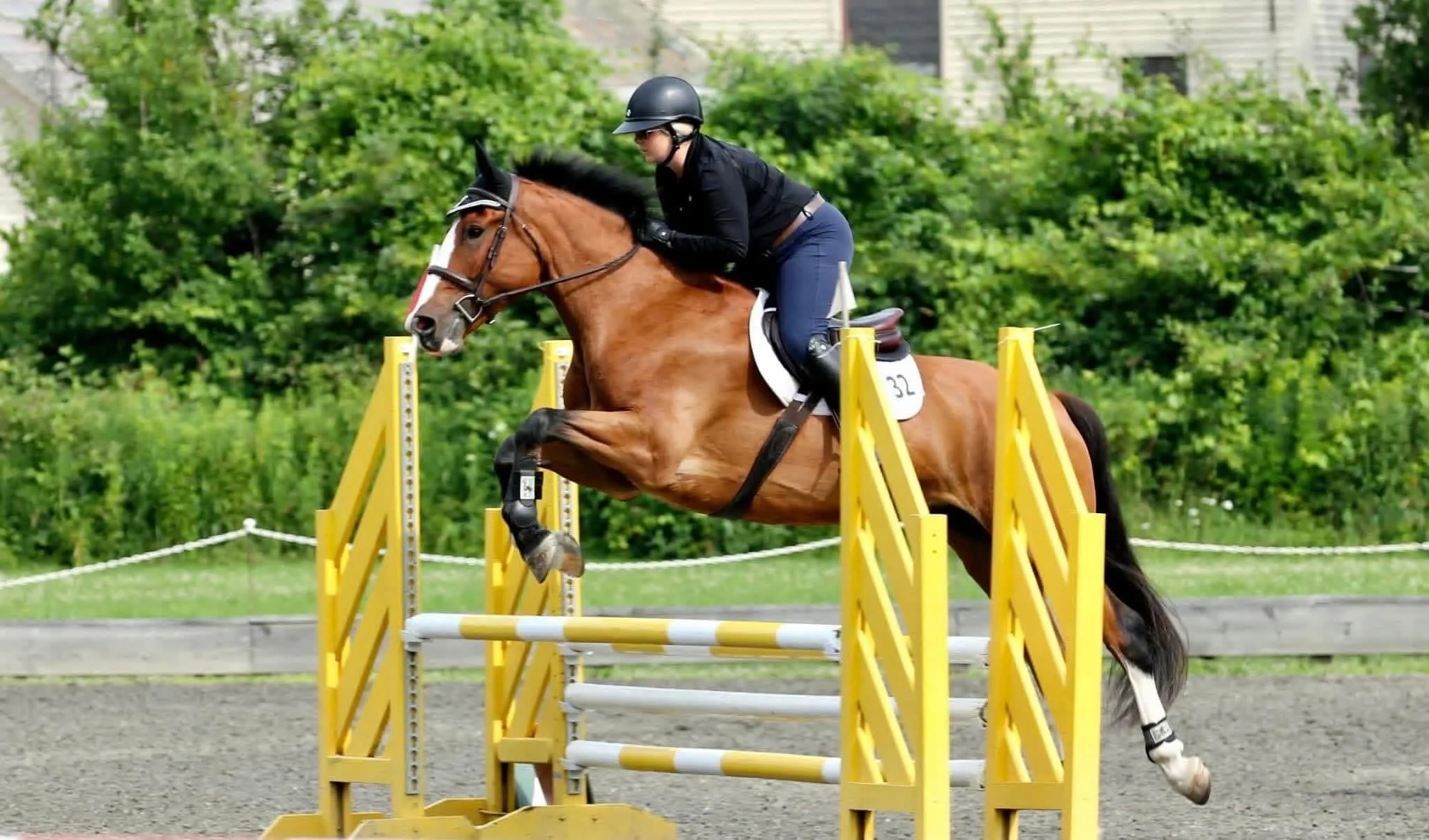 A person wearing a helmet and black riding jacket riding a brown horse jumps over a wooden obstacle during an equestrian show on a sunny day.