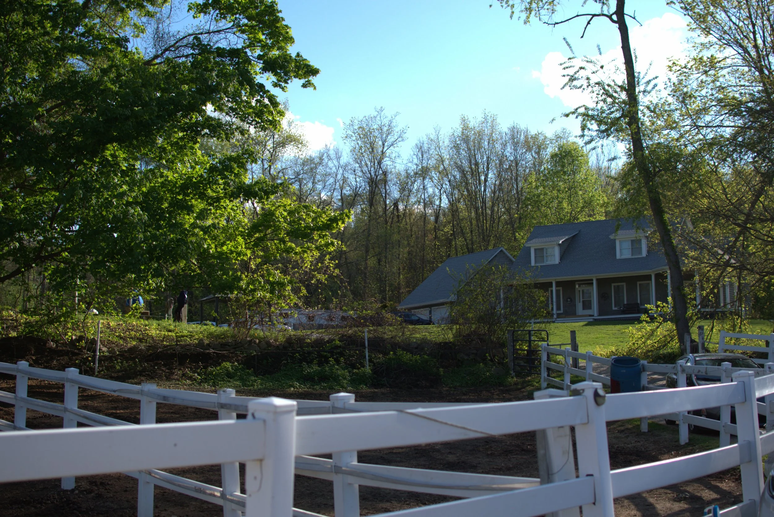 An outdoor scene with a white wooden fence in the foreground, a garden patch with soil behind it, and a house with a gray roof and white windows in the background. There are lush green trees and a partly cloudy blue sky overhead.
