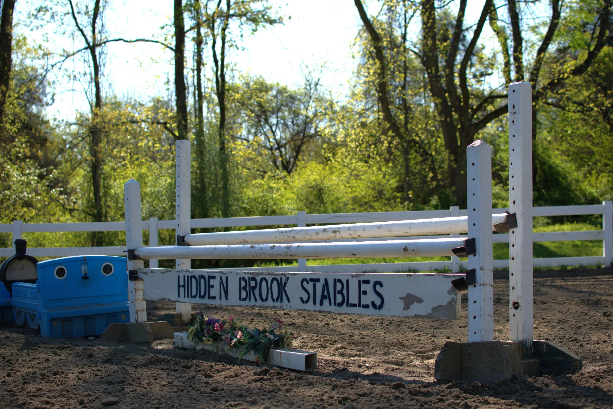 Show jumping obstacle at Hidden Brook Stables with a white rail and a sign, surrounded by dirt and some flowers, with green trees in the background