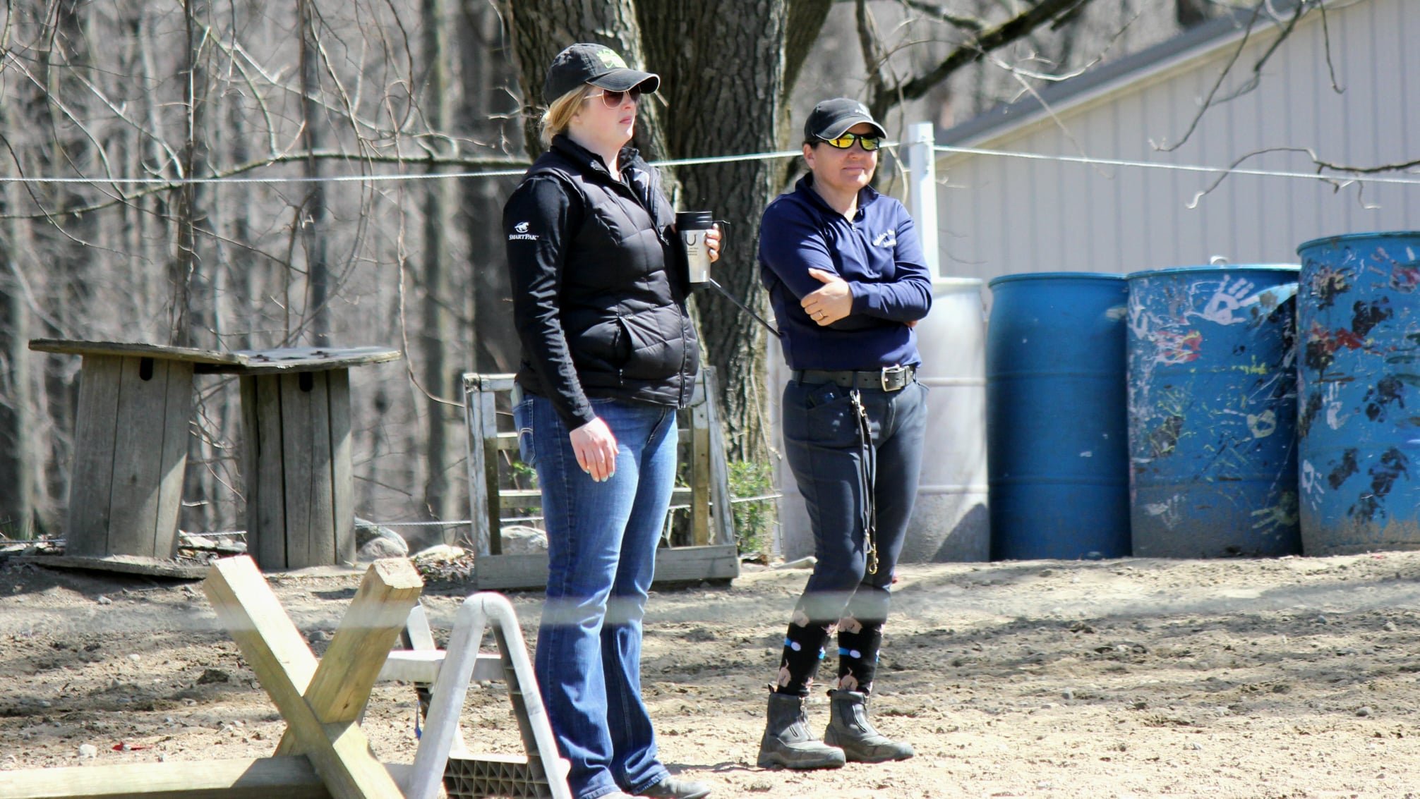 Two women standing outdoors near trees, one holding a travel mug, the other with arms crossed, with large blue barrels and wooden structures in the background.