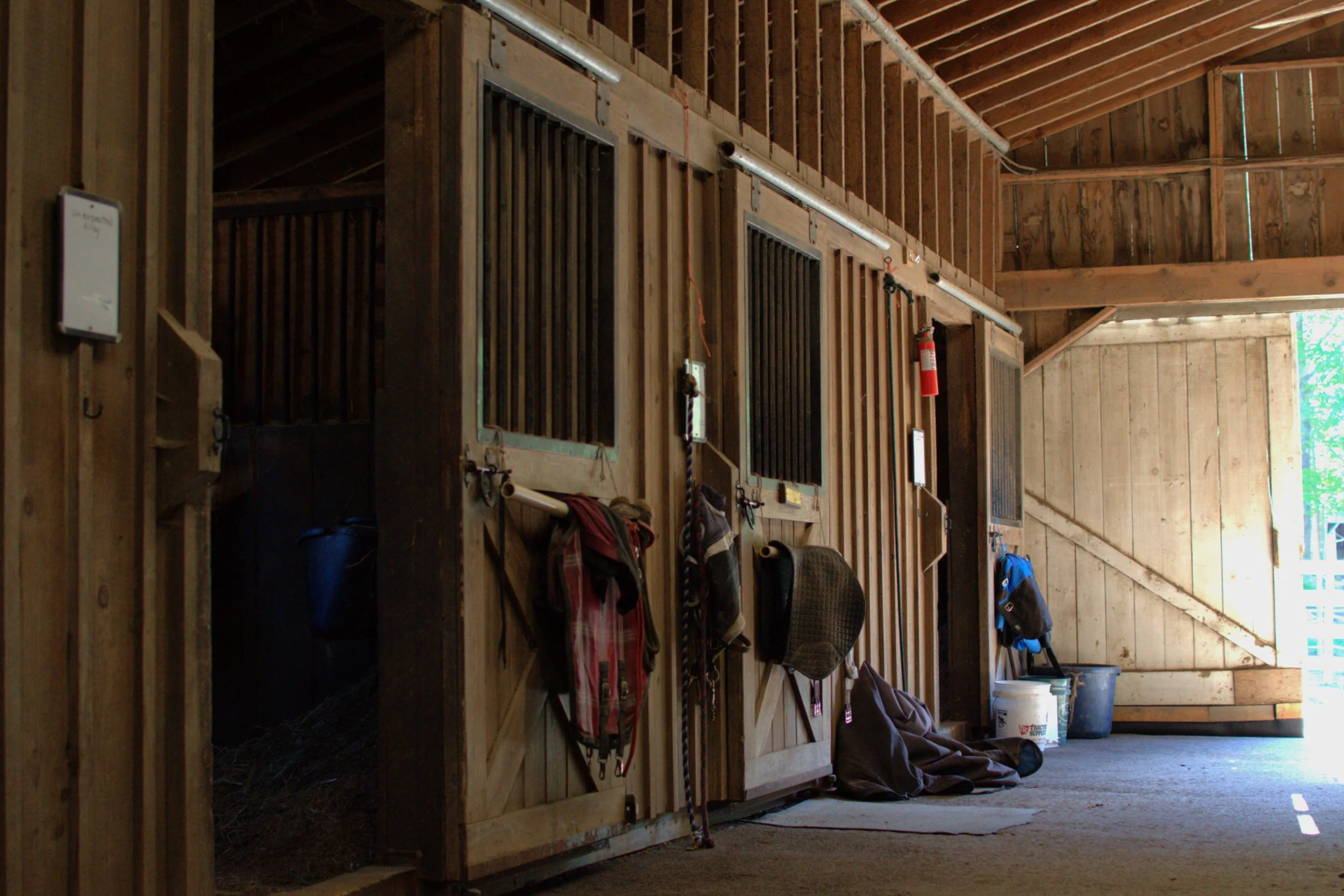 Interior of a barn with wooden walls, hooks, and storage items, with natural light coming from an opening at the end.