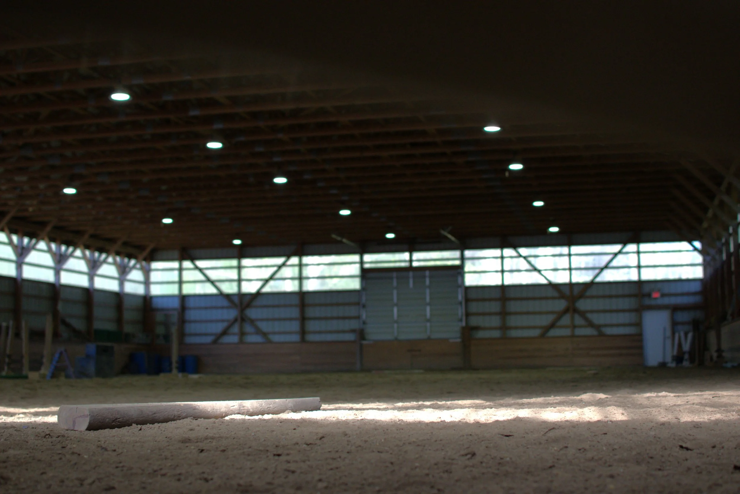 Interior of an empty indoor riding arena with dirt floor, wooden beams supporting the ceiling, and windows allowing natural light.
