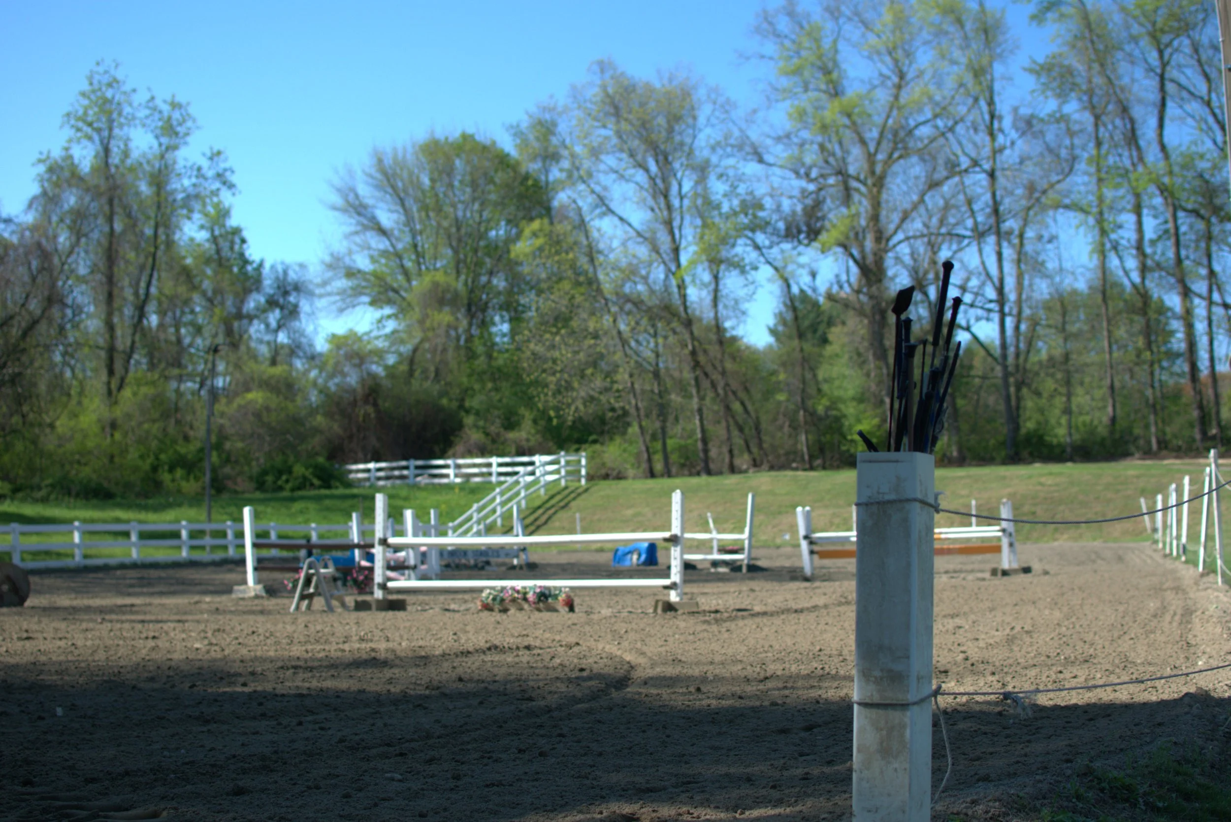 An outdoor horse riding arena with jumps, surrounded by white fencing and trees in the background, under a clear blue sky.