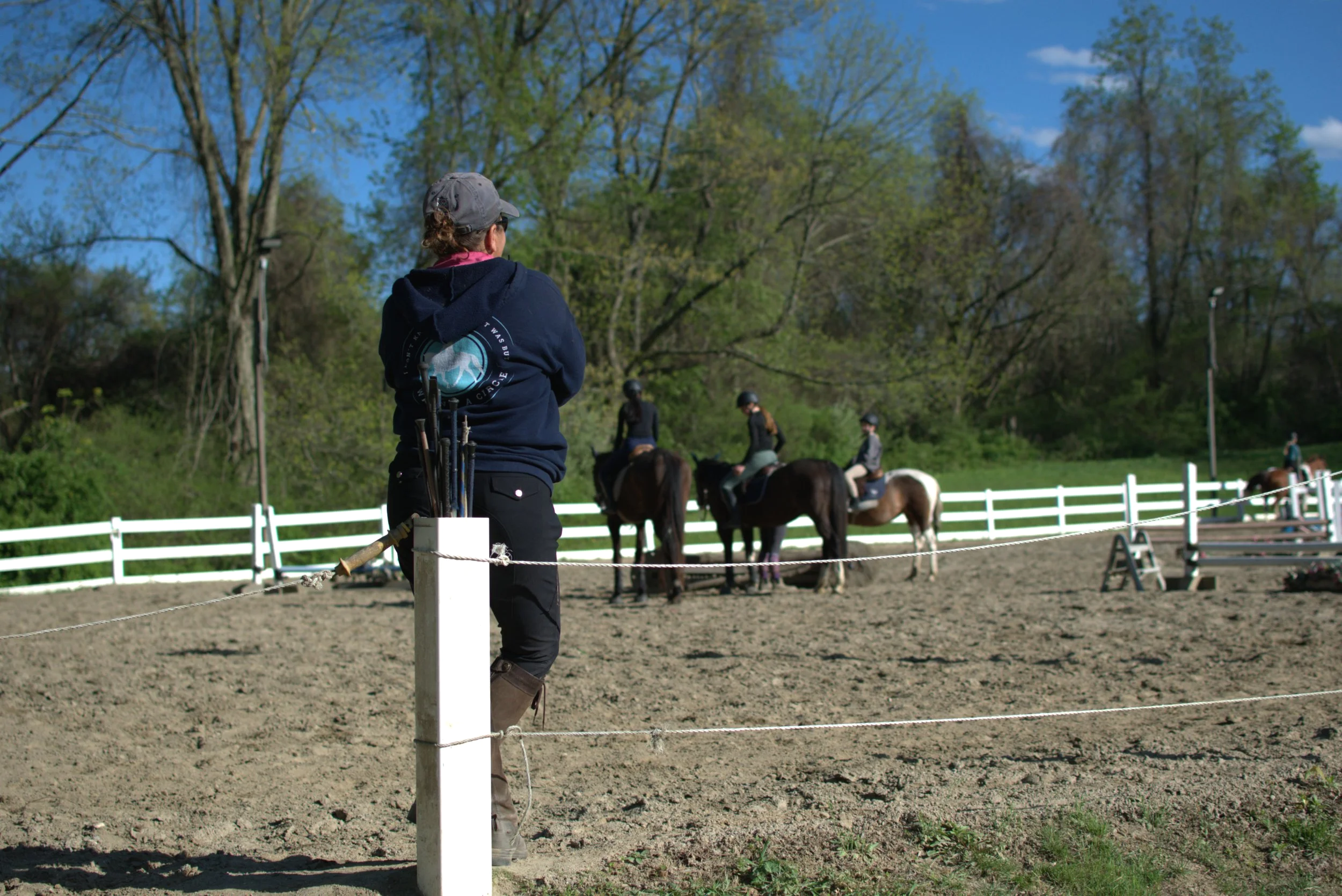 A person with a hat and dark hoodie standing by an outdoor riding arena, observing horseback riders and trainers. The arena has a white fence, and there are trees and a blue sky in the background.