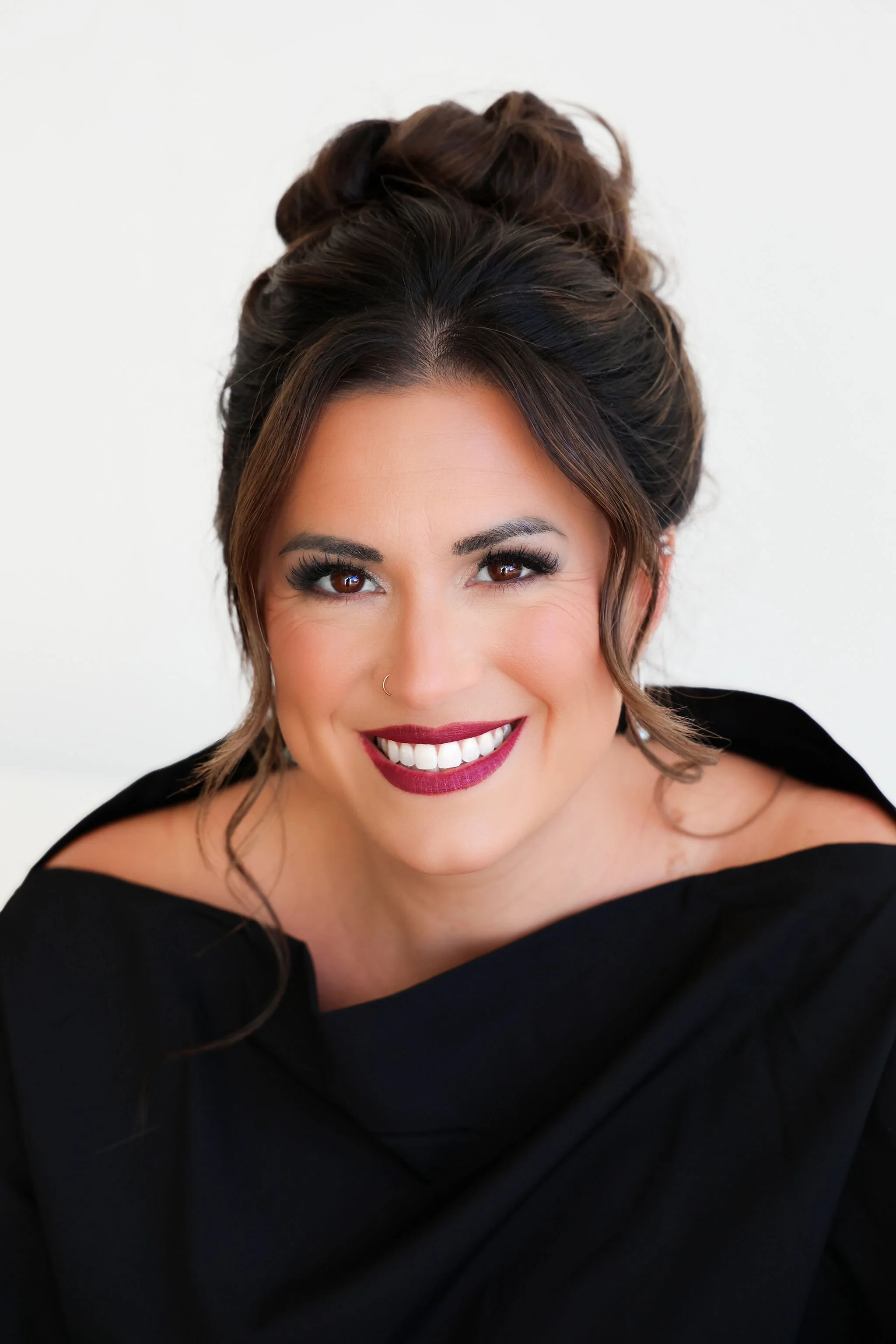 Close-up of a smiling woman, Danielle Larish, with brown hair styled in an updo, wearing makeup including dark lipstick and eye makeup, and a black off-the-shoulder top against a plain white background.