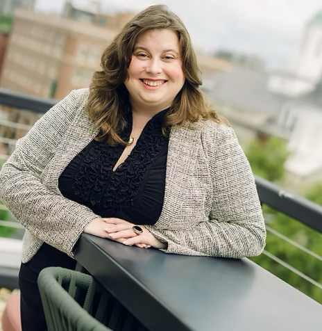 Joyce Deuley, a woman with brown hair smiling on rooftop balcony, wearing a gray blazer and black top, with a cityscape in the background.