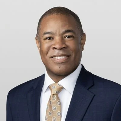 Professional headshot of Ronald D. Sullivan, a man wearing a navy blue suit, white shirt, and a light-colored patterned tie, smiling at the camera.