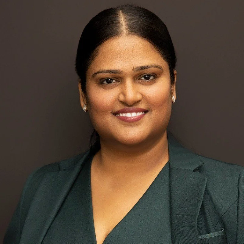 Professional portrait of, Niti Vanee, a woman with dark hair in a bun, wearing pearl earrings and a dark green blazer, smiling at the camera against a dark gray background.