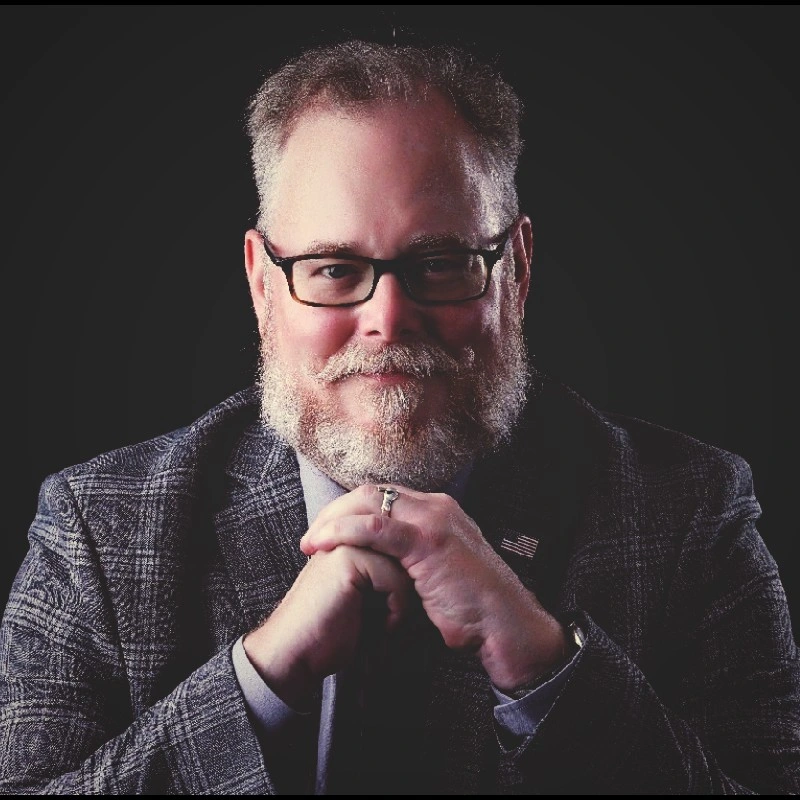 A man, Art Trevethan with glasses, a beard, and gray hair in a checkered blazer, sitting with hands clasped, smiling in a professional portrait against a black background.