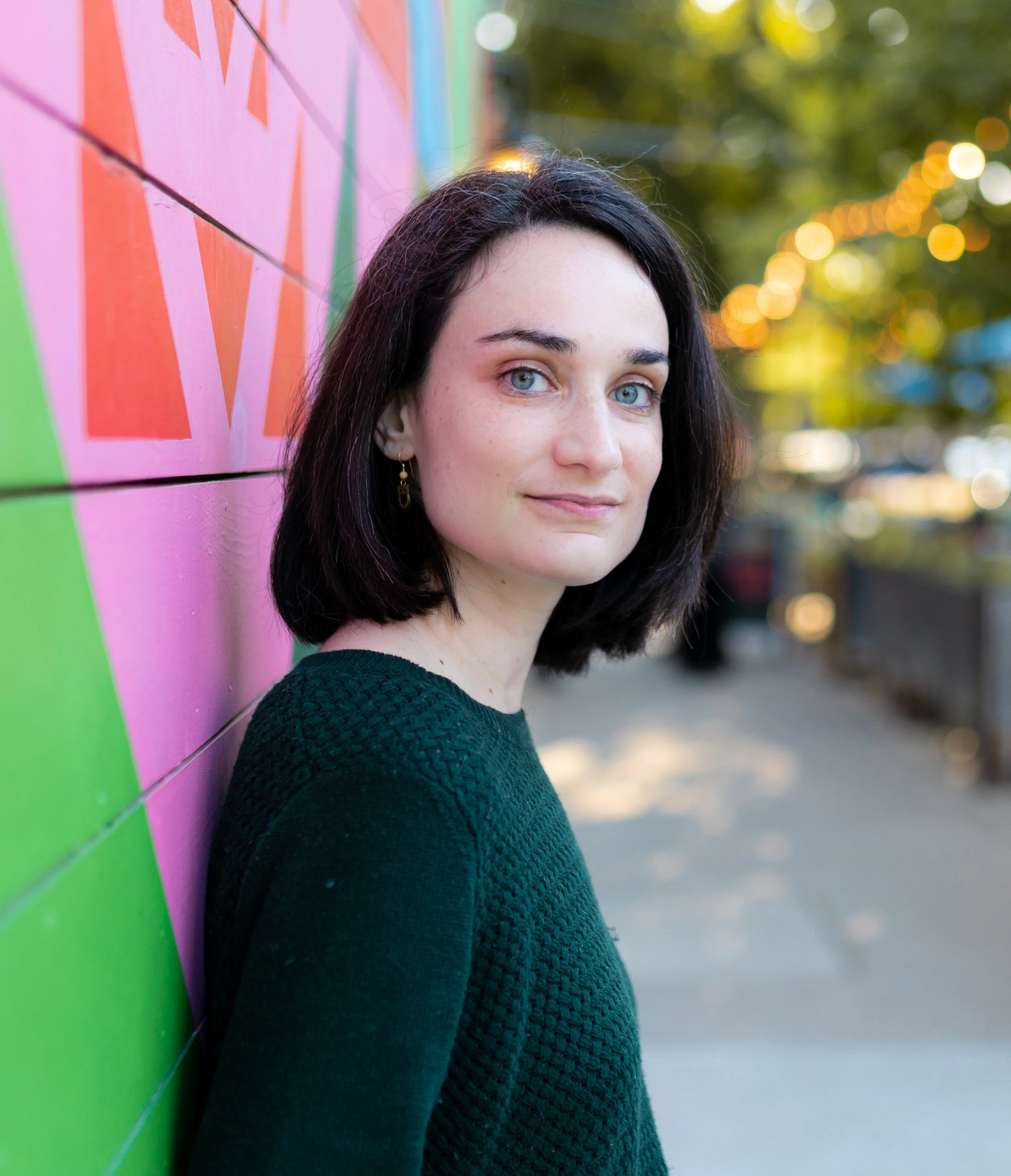 A young woman with dark hair and blue eyes stands near a colorful painted wall, with blurred outdoor lights and trees in the background.