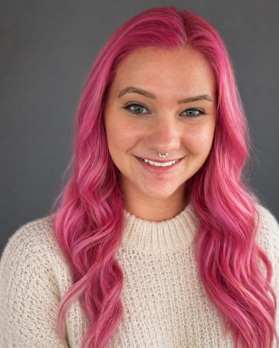 A woman with long, wavy pink hair and blue eyes smiling at the camera with a septum piercing, wearing a cream-colored knit sweater against a dark gray background.