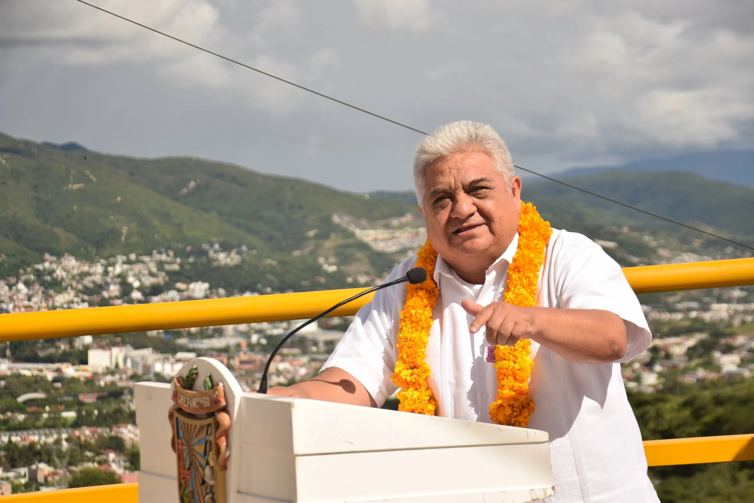 A man with white hair wearing a white shirt and a yellow flower garland, speaking into a microphone on a podium with a colorful pattern, outdoors with hills and a cloudy sky in the background.