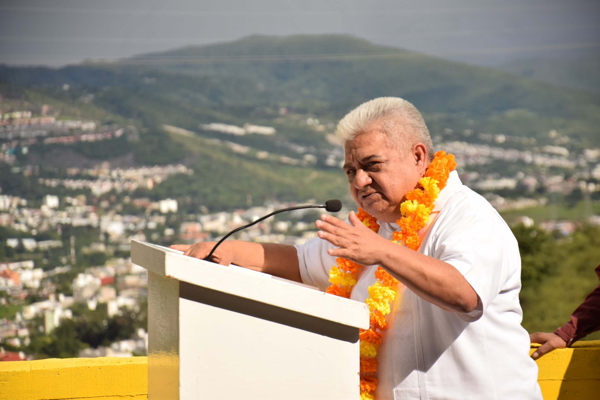A man with gray hair wearing a white shirt and a marigold flower garland speaking at a podium during an outdoor event, with a hilly cityscape and mountains in the background.