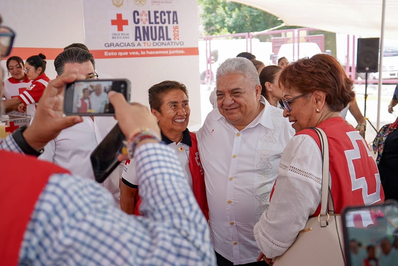 People smiling at an event with Red Cross banners and volunteers. A woman in a black shirt and man in white shirt hug, with others taking photos.