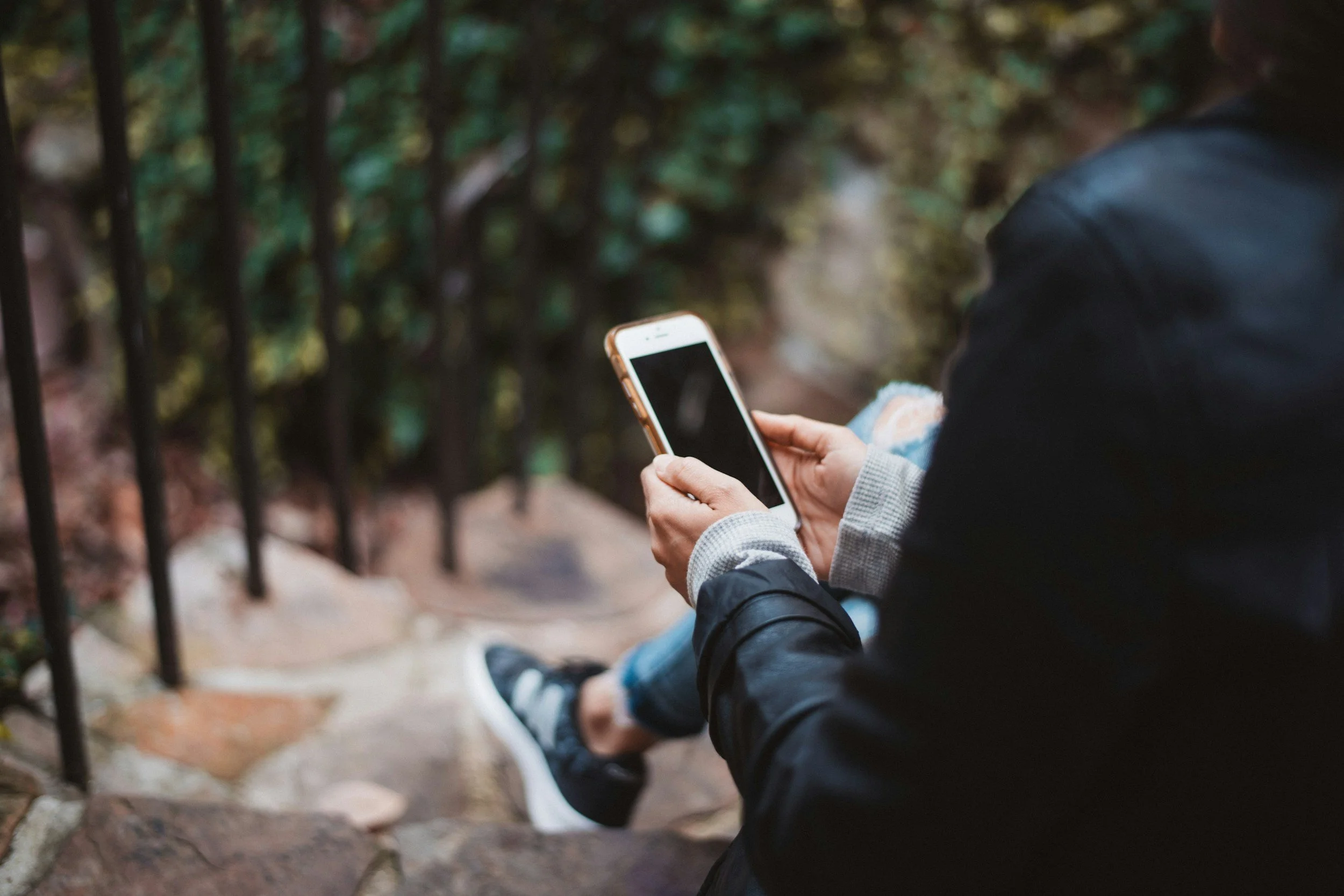Person in dark jacket and gloves holding a smartphone, sitting on stone steps near a metal fence, with green bushes in the background.