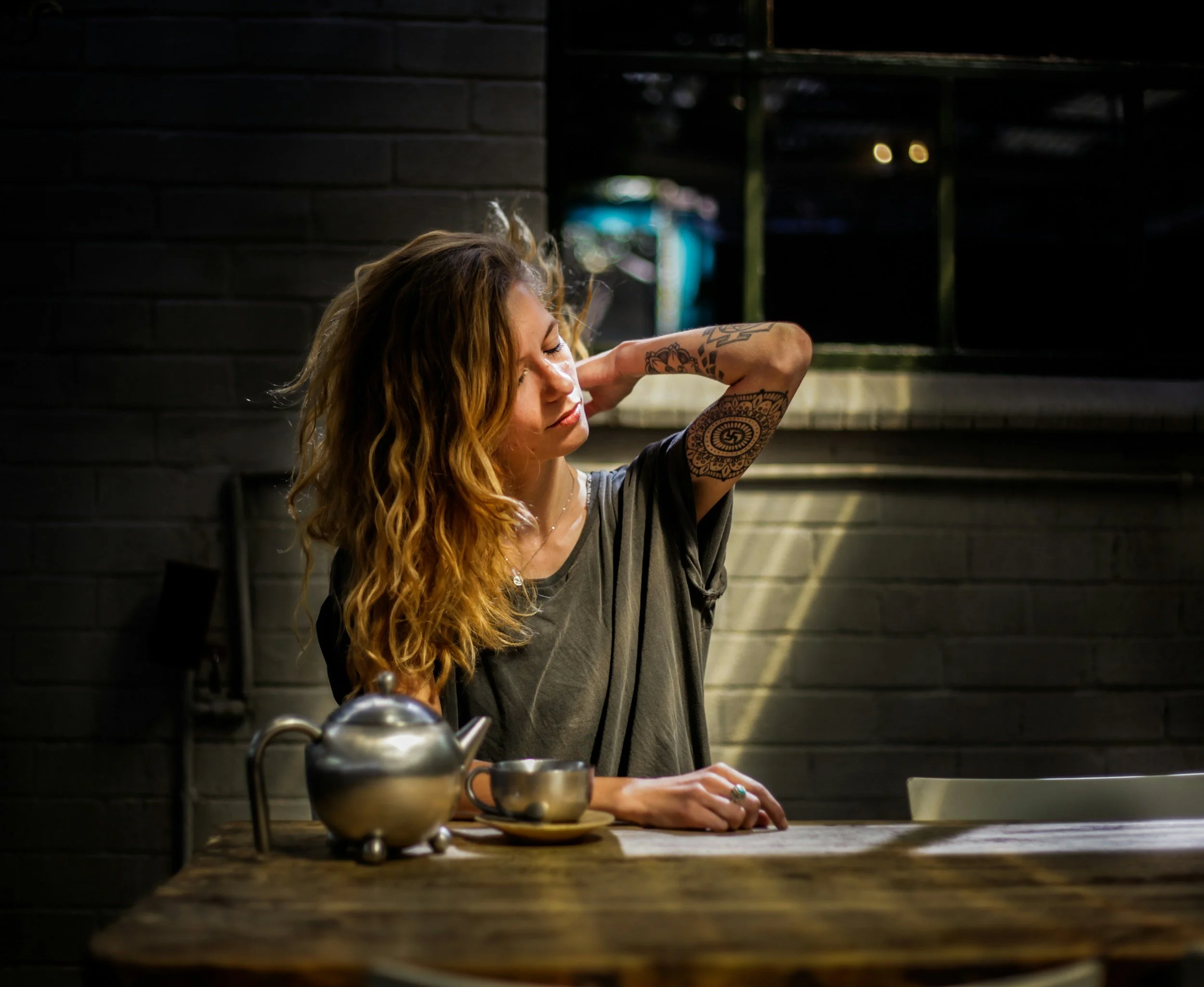 A woman with long, curly hair and tattoos on her arm sits at a wooden table in a dimly lit room, with her eyes closed and one arm resting behind her head. There are teapots or kettles on the table, and the background features a black brick wall with a window showing some lights outside.