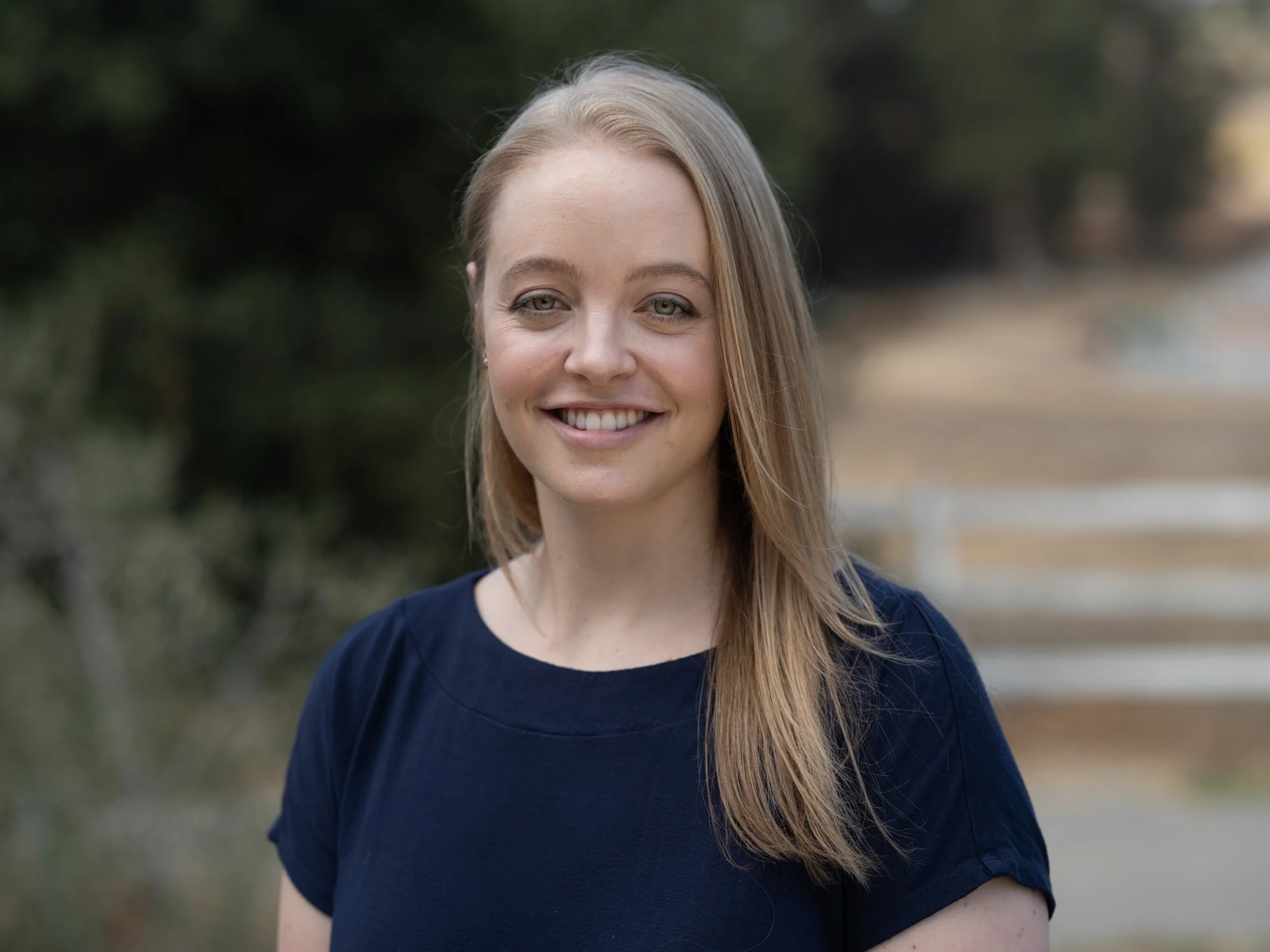 A young woman with long blonde hair, light green eyes, and fair skin, smiling outdoors near trees.