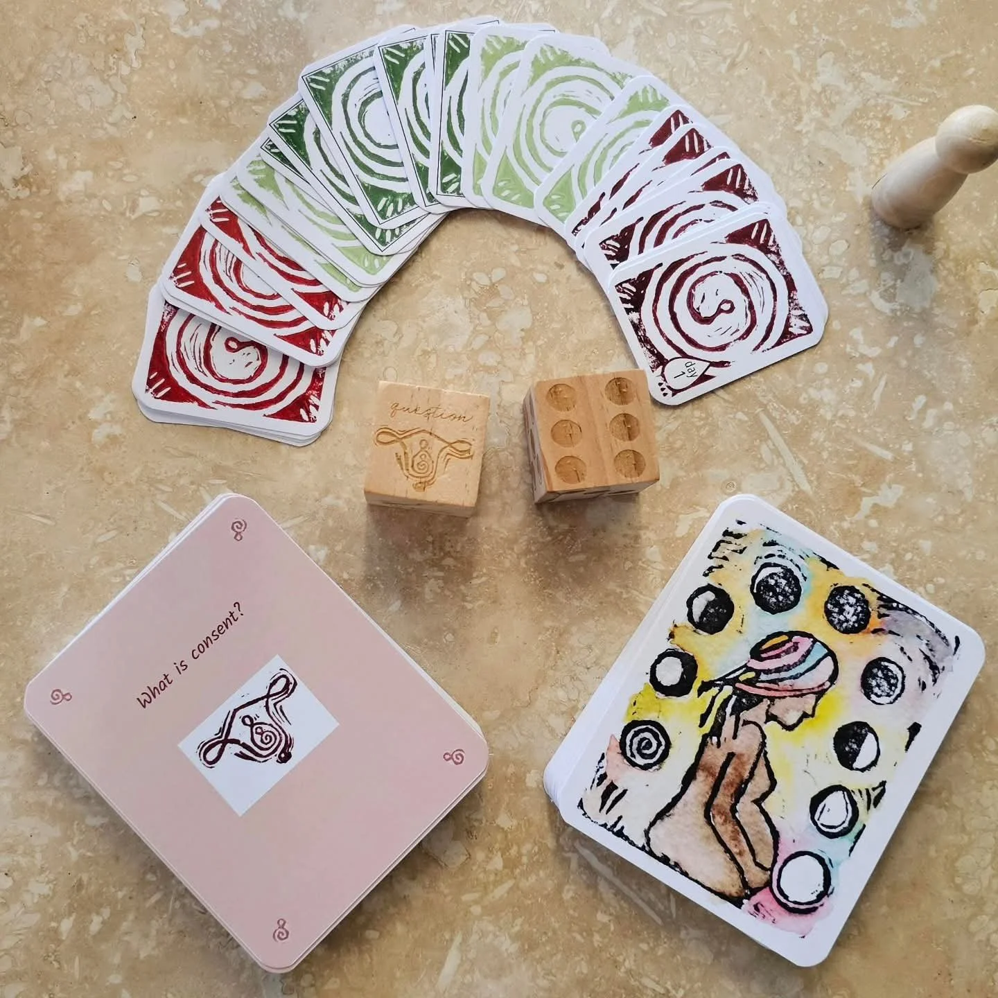 Know Your Flow game pieces on a stone table: a fan of red and green hand-printed spiral cycle cards, a wooden question die and a number die, a pink question card reading “What is consent?”, a watercolor moon-phase art card, and a wooden player token.