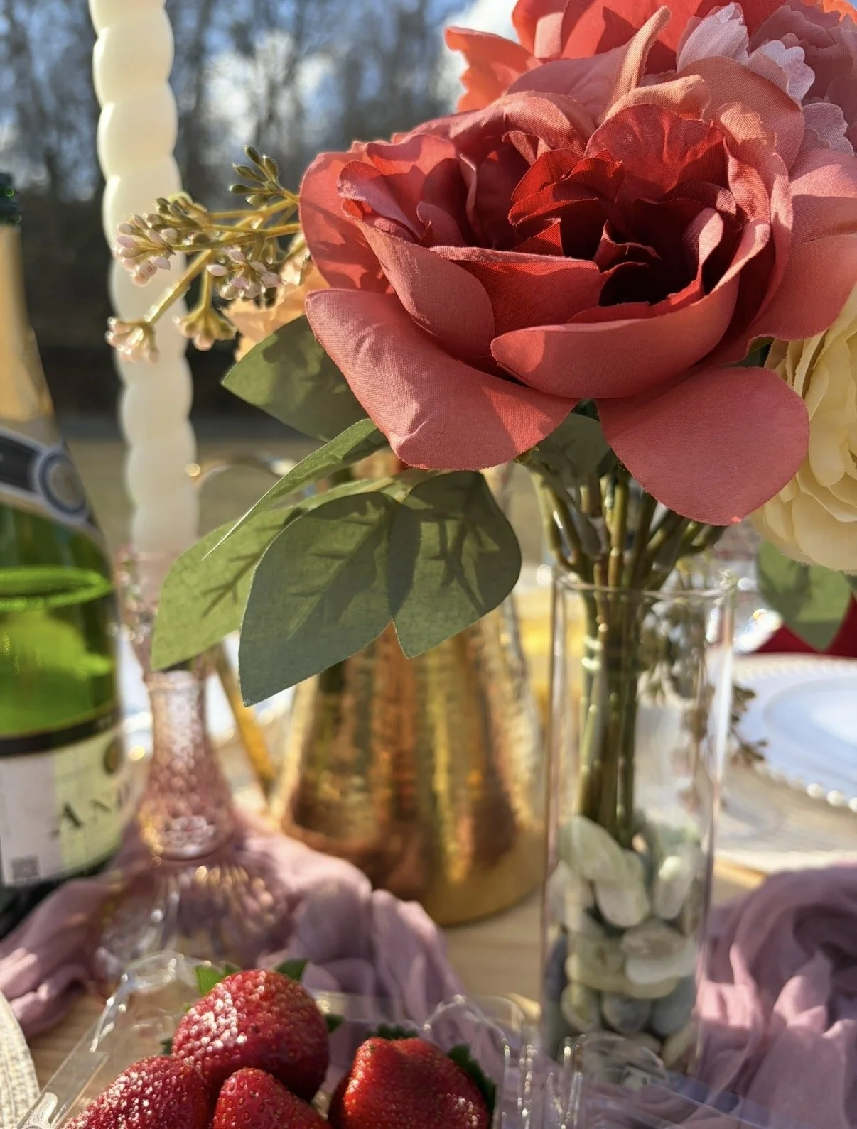 Close-up of a bouquet of pink and cream artificial flowers in a glass vase, with strawberries in a container in the foreground. The scene is illuminated by sunlight.