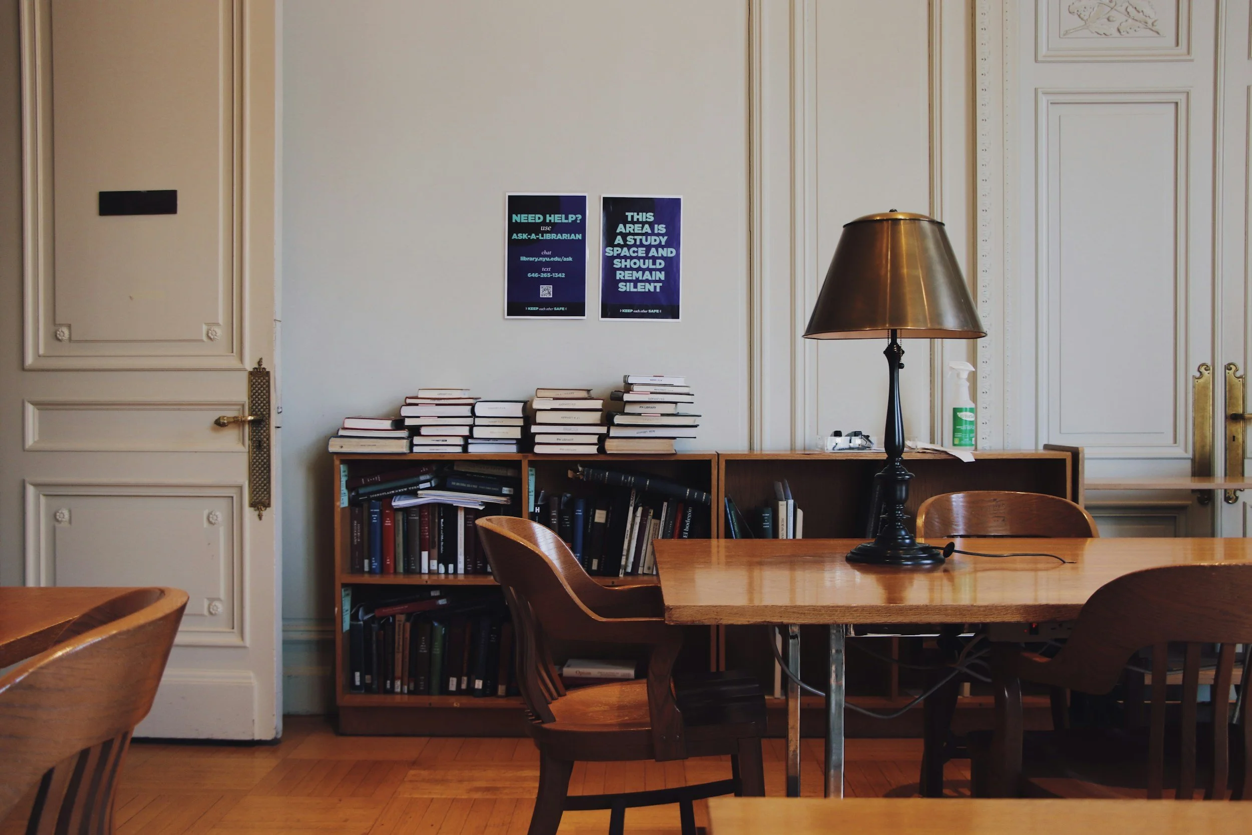 A study room with wooden tables and chairs, a tall wooden bookshelf filled with books, two informational posters on the wall, a black table lamp, and a bottle of cleaning spray on the desk.