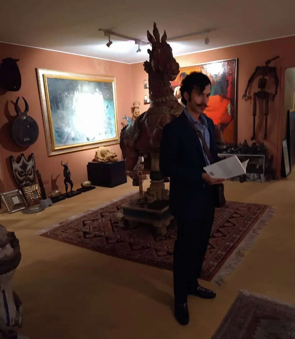 A man dressed in a suit with a mustache holding a book, standing in a room decorated with various sculptures and artworks, including a large wooden horse sculpture and a skeleton, surrounded by paintings and artifacts.