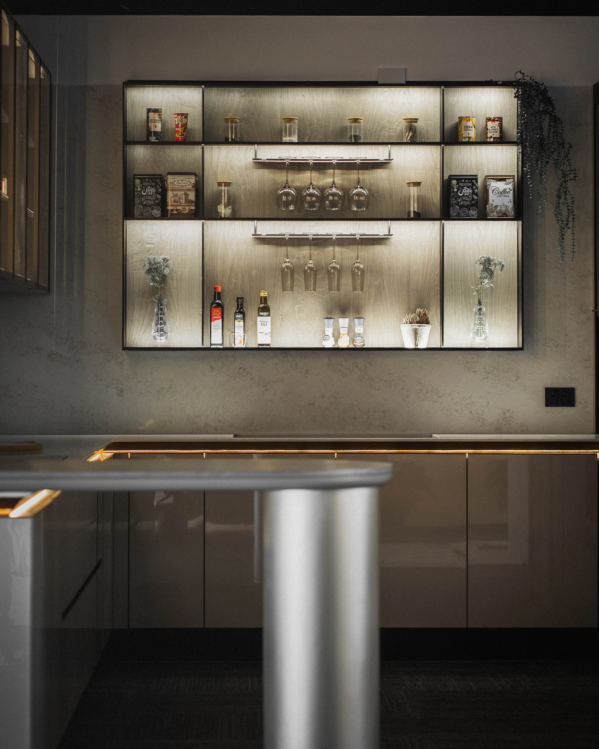 A modern, dimly lit kitchen display shelf with glassware, coffee books, and decorative items, seen from below with a black ceiling and wall in the background.