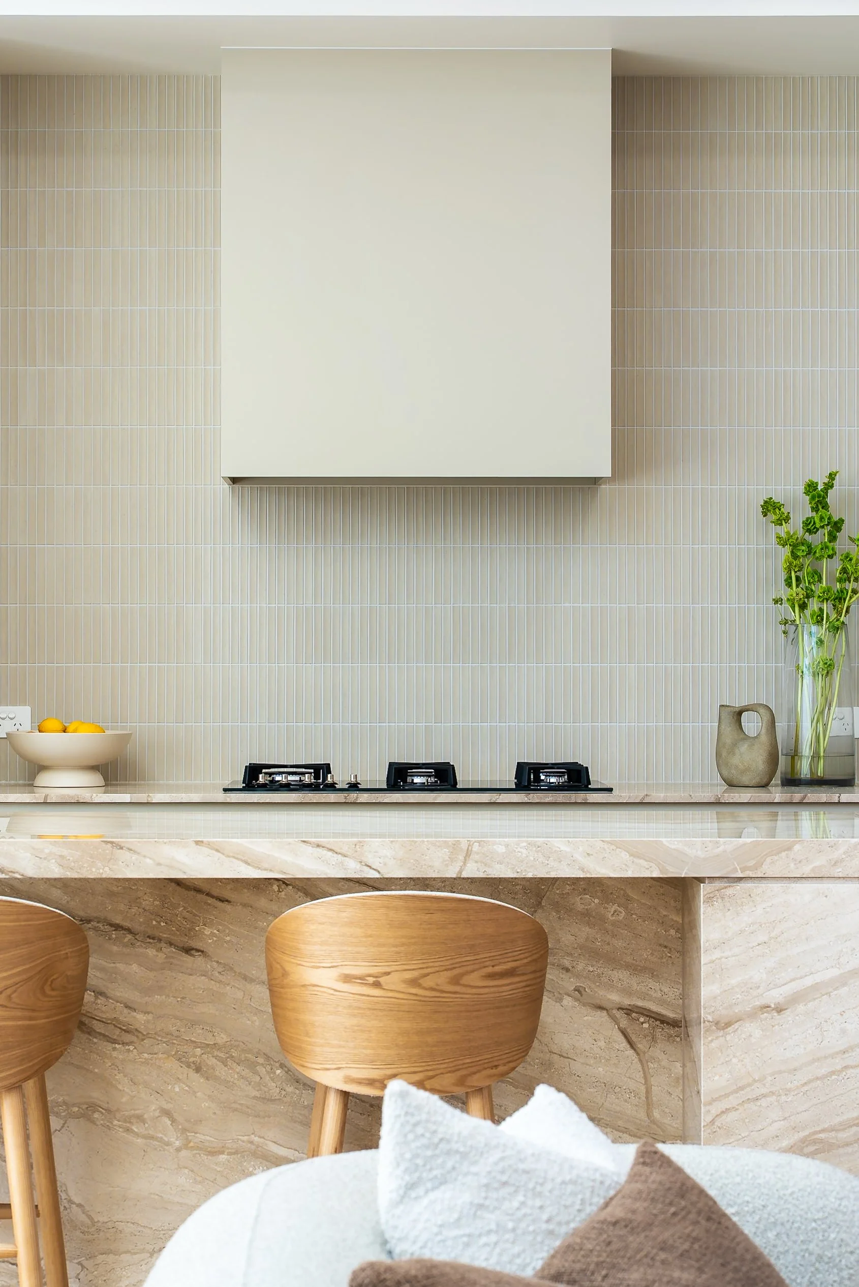 Modern kitchen with beige and white tiled walls, a white range hood, a marble countertop, wooden stools, a white bowl with lemons, a plant, and a decorative vase.