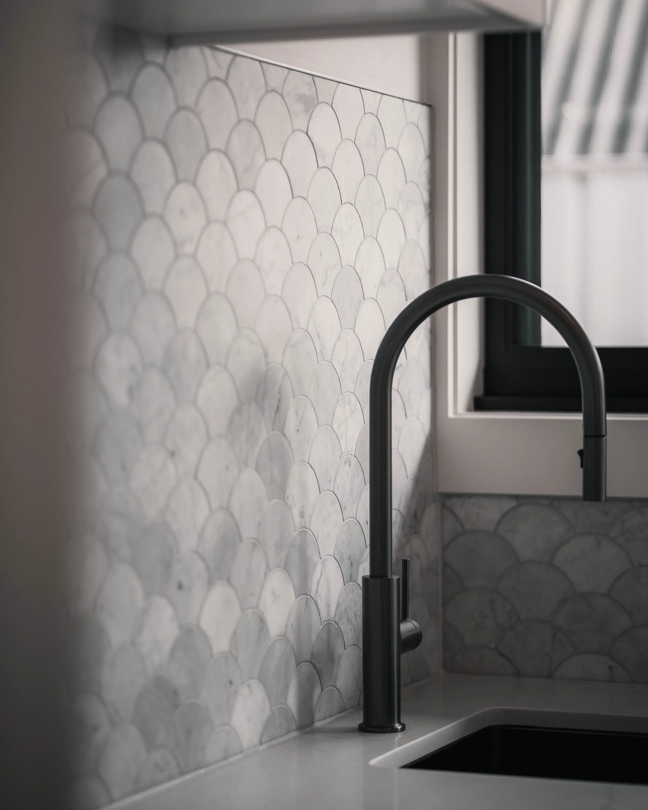Close-up of a kitchen countertop with a modern black faucet, a textured white tile backsplash with a fish-scale pattern, and a window with black framing in the background.