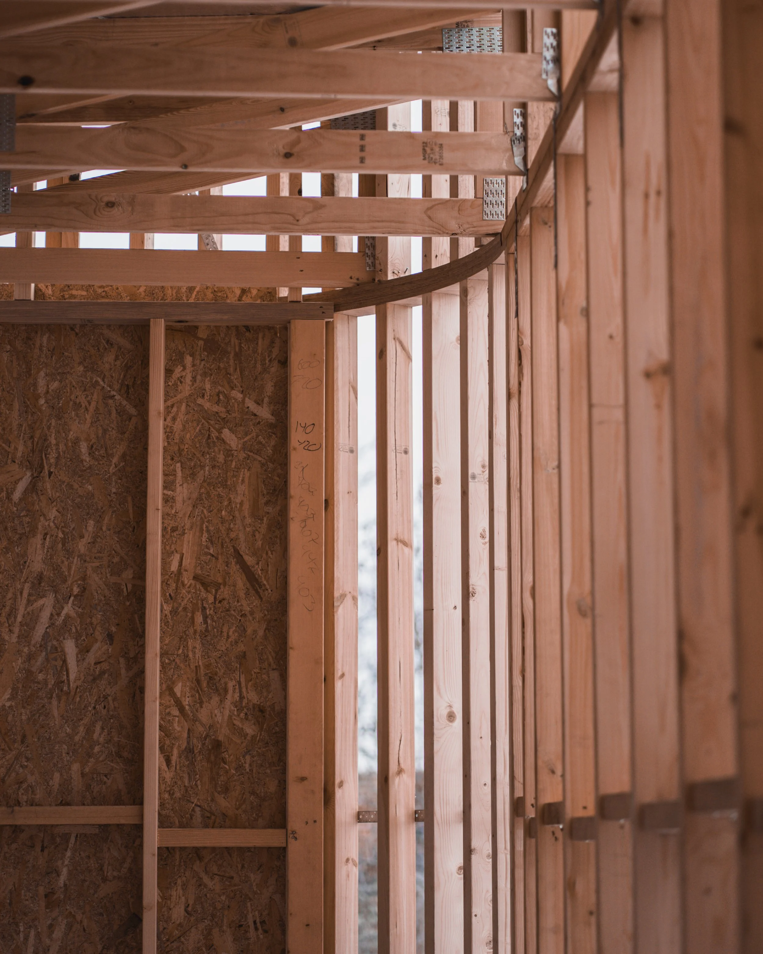 Interior view of a wooden house under construction showing vertical wooden studs and metal connectors for wall framing, with some markings written on the wood.