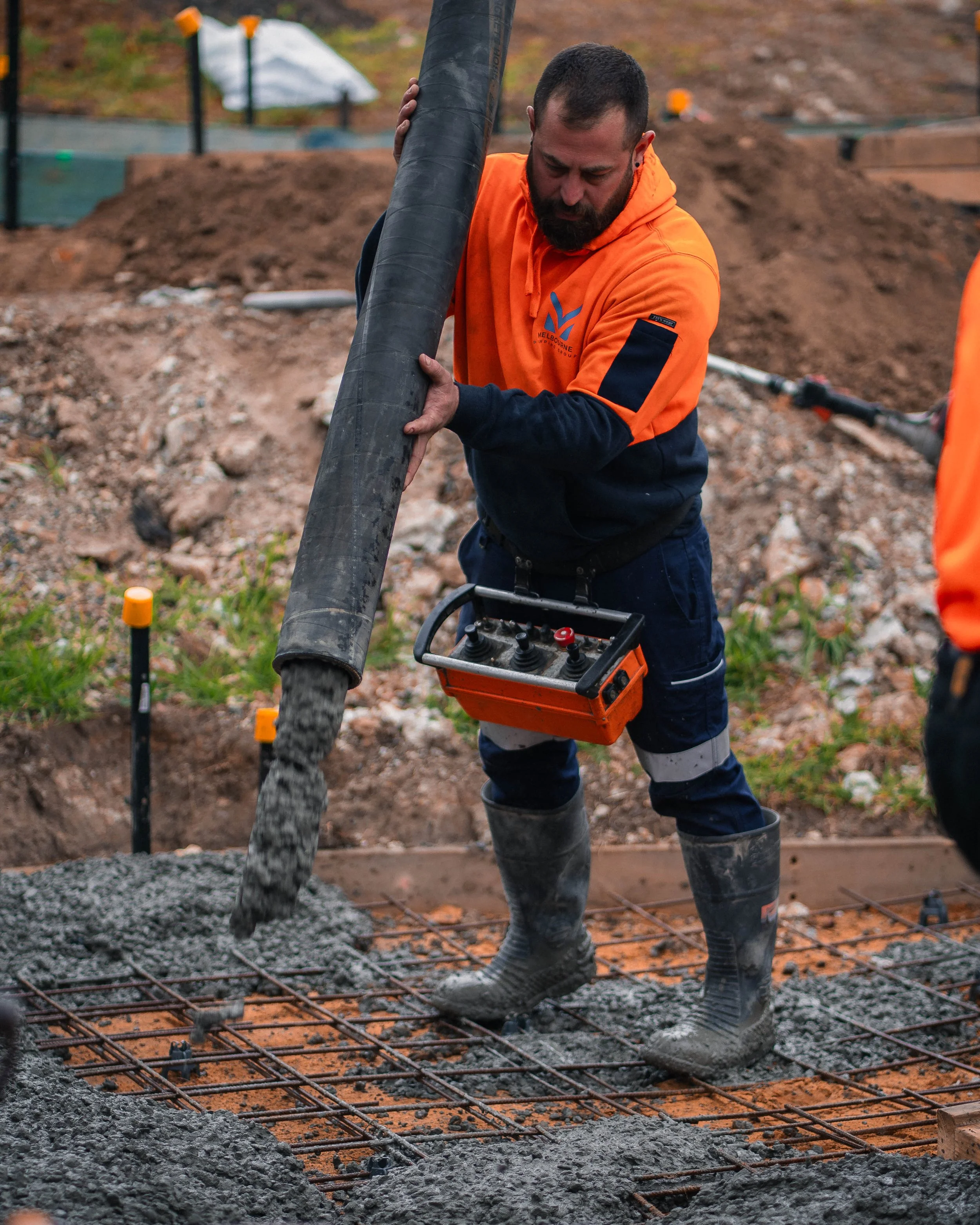 A construction worker pouring concrete over a rebar grid at a construction site. The worker is wearing an orange hoodie, navy gloves, and knee-high boots, and is using a machine to spread the concrete.