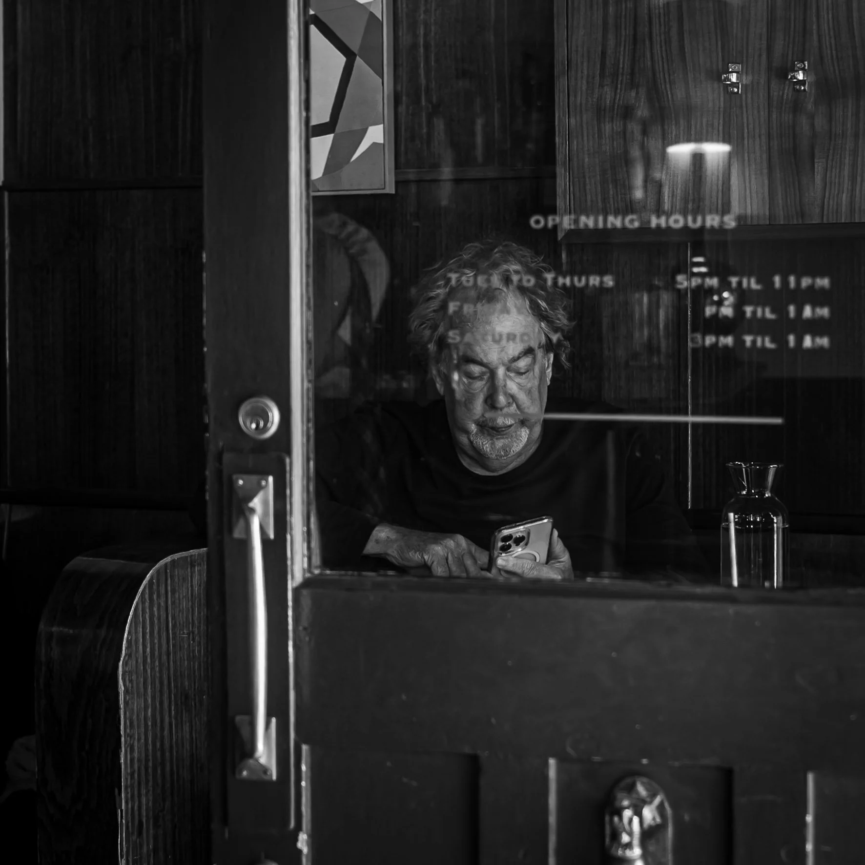 An older man with glasses and gray hair sitting at a table in a restaurant or cafe, looking at a smartphone, with a reflective glass window with text about the establishment's opening hours.