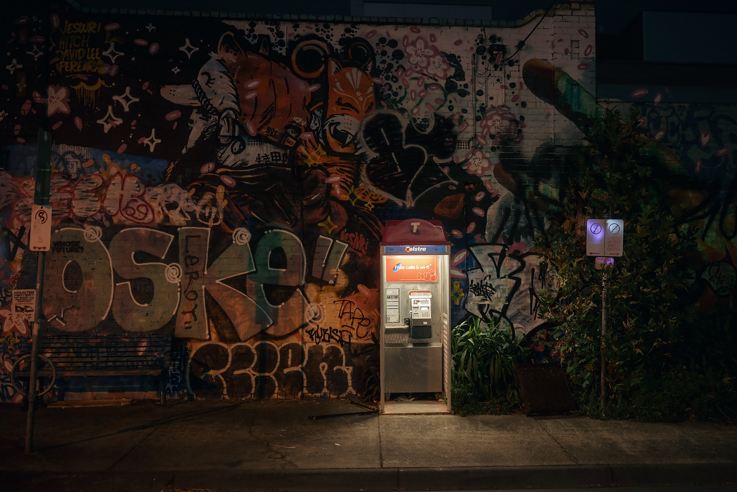 Nighttime street art mural featuring two men in combat gear surrounded by flowers and stars, painted on a brick wall, with a payphone booth and some greenery in front.