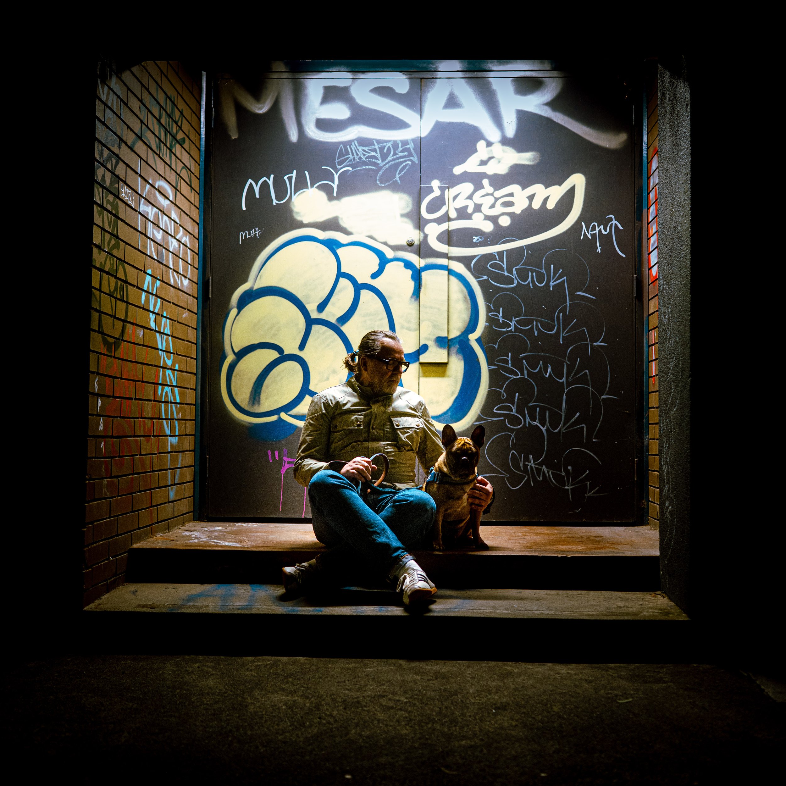 A man with glasses and a beard sits on the ground with a dog, against a background of a brightly lit graffiti-covered metal door, framed by brick walls in a dark alleyway.