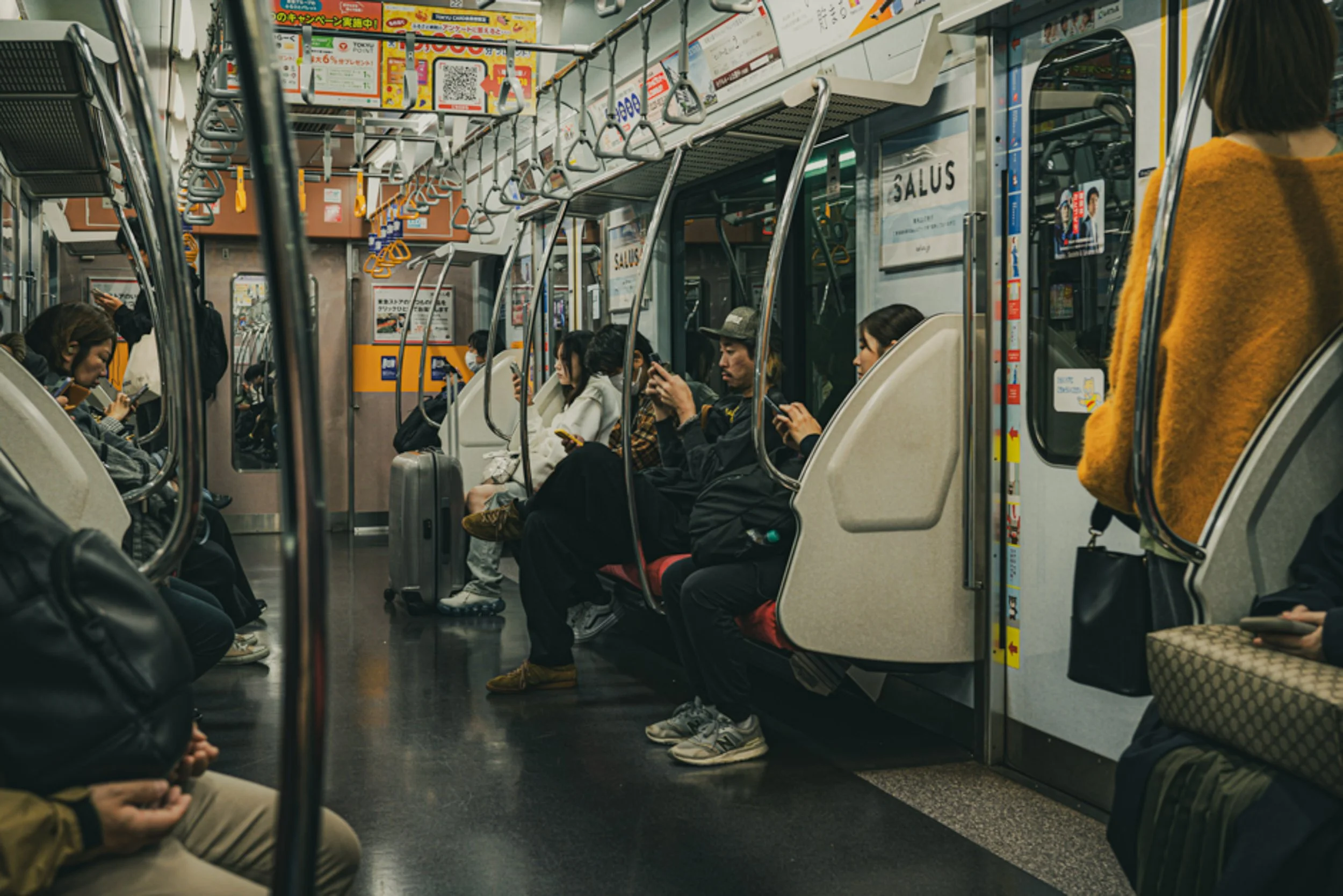 People sitting and standing inside a subway train, some are using their phones, and advertisements are visible on the walls.