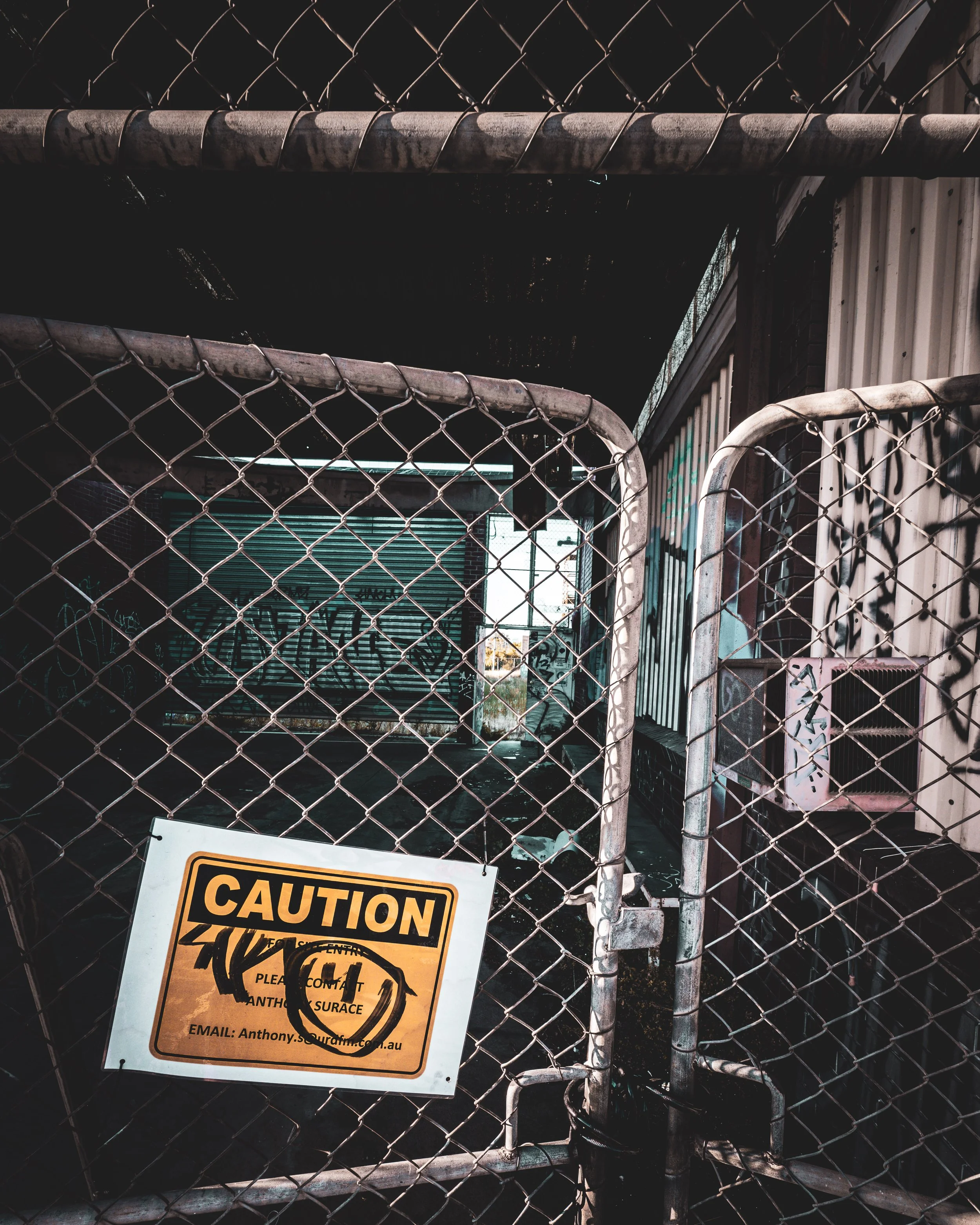 A chain-link fence with a caution sign attached, warning about entering a restricted area. The fence surrounds what appears to be an industrial or construction site with metal structures and a partially visible background.