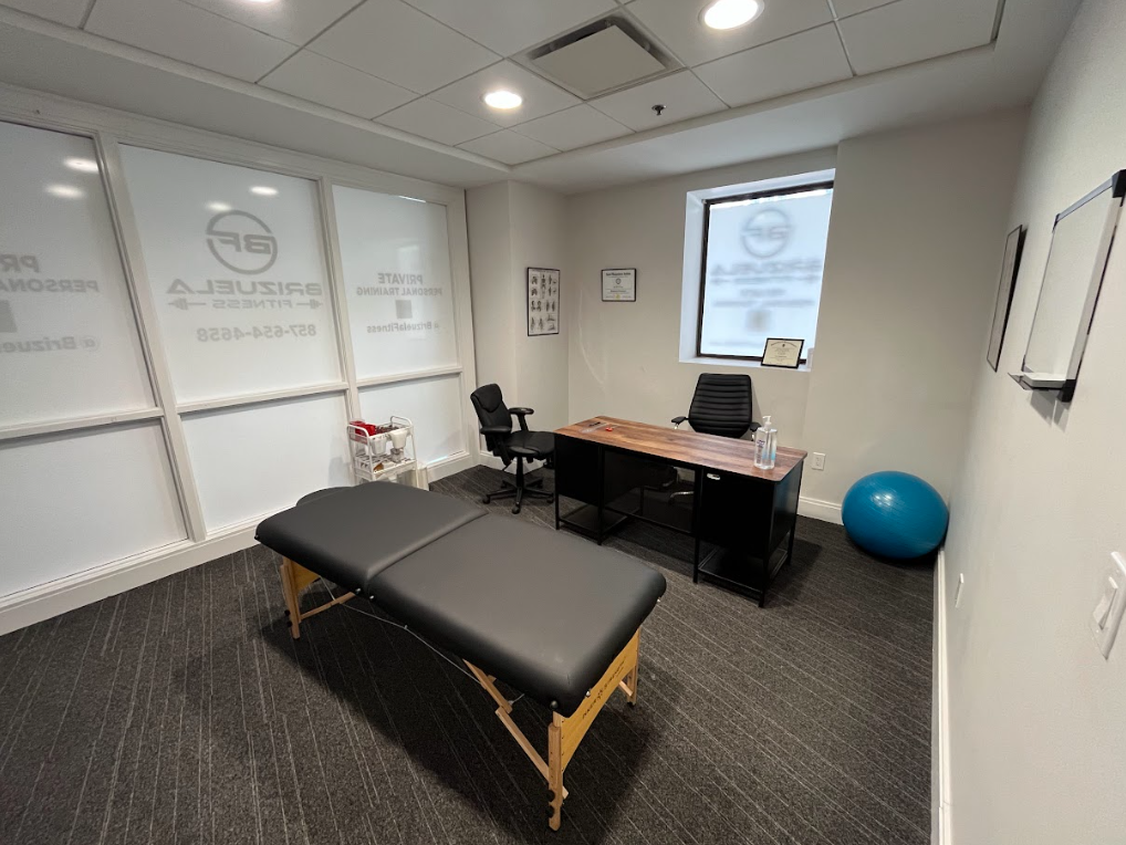 Empty massage therapy or physical therapy office with treatment table, desk, two chairs, exercise ball, and framed certificates on the wall.