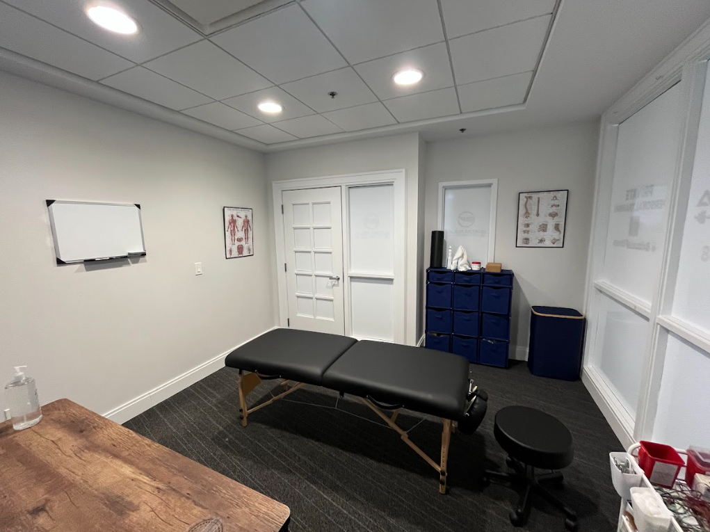 Interior of a massage therapy or chiropractic treatment room with a massage table, medical charts, and storage cabinets.