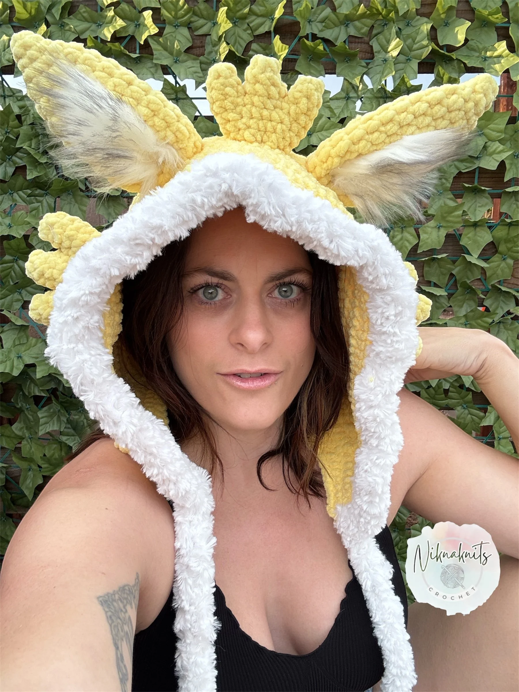 A woman taking a selfie wearing a yellow plush reindeer hat with antlers and ears, and a white furry trim, outdoors with a green leafy background.