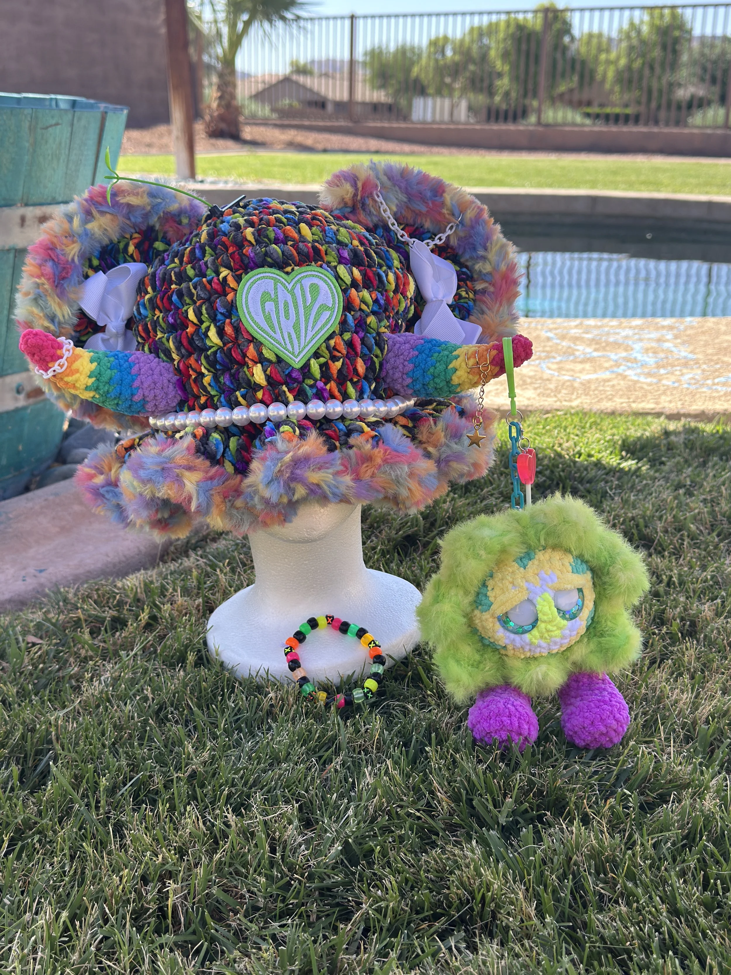 Colorful plush stuffed animal with rainbow fur, beads, and charms, placed on a white stand outdoors with a bracelet and a colorful plush toy nearby, grass, a pool, and a backyard fence in the background.