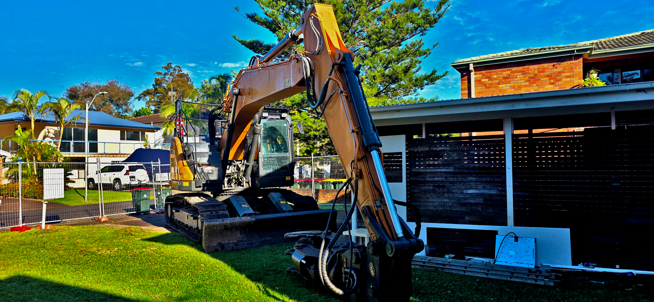 A construction excavator parked on a grassy yard in front of a house with a brick exterior, with trees and other houses visible in the background.