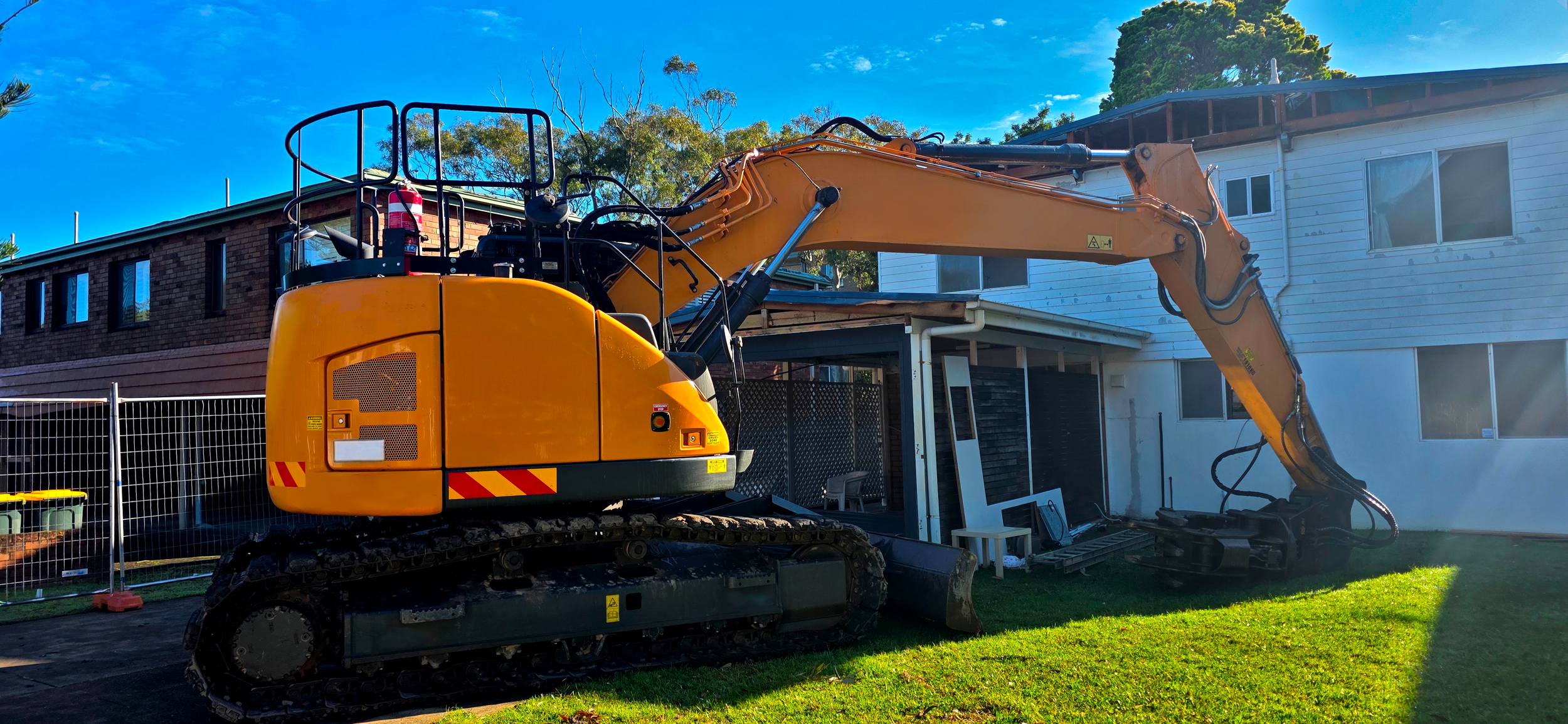 A yellow excavator is positioned in a backyard, with a house and trees in the background, under a clear blue sky.