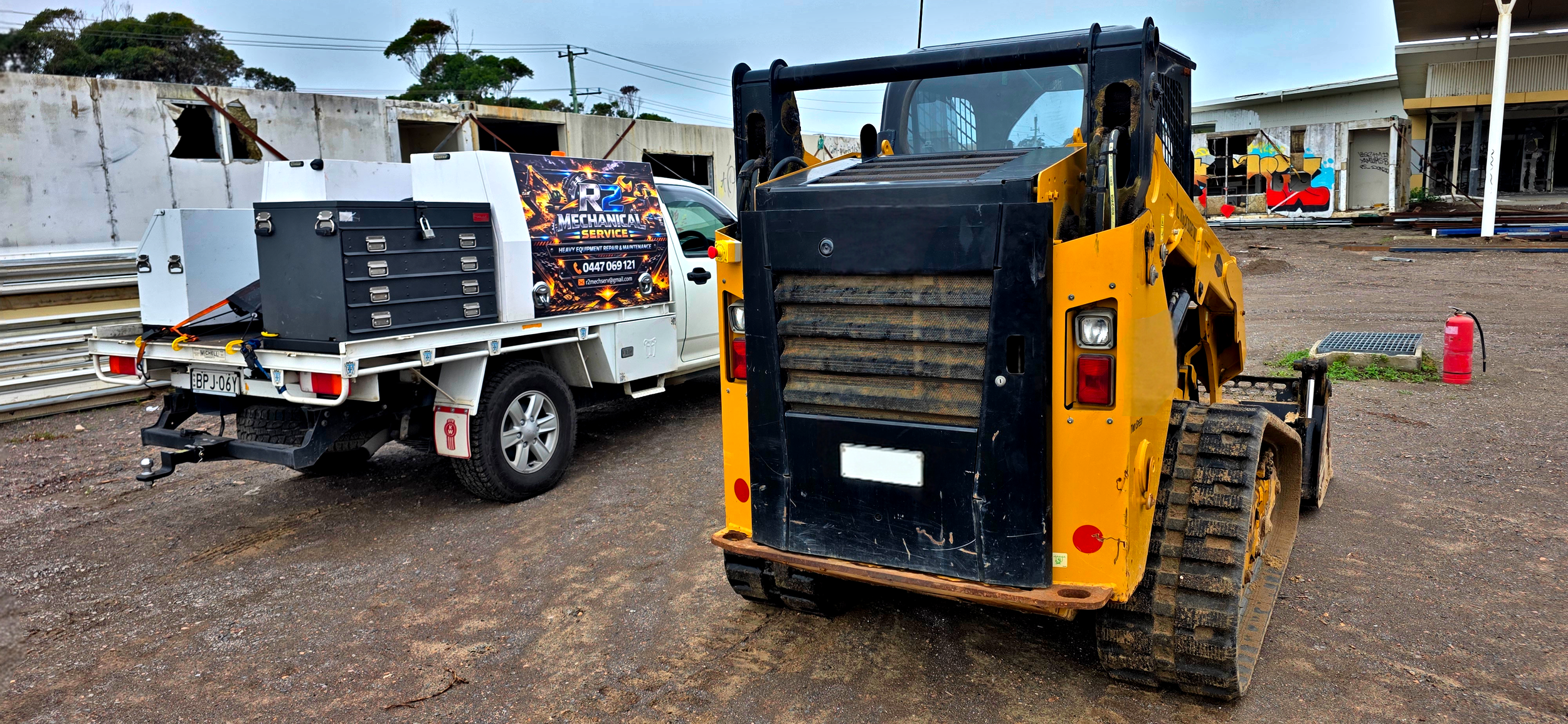 A construction site with a yellow skid-steer loader and a white utility truck. The truck has a sign advertising heavy equipment repair service, and tools are loaded onto it. The background features a partially constructed building and graffiti on a w