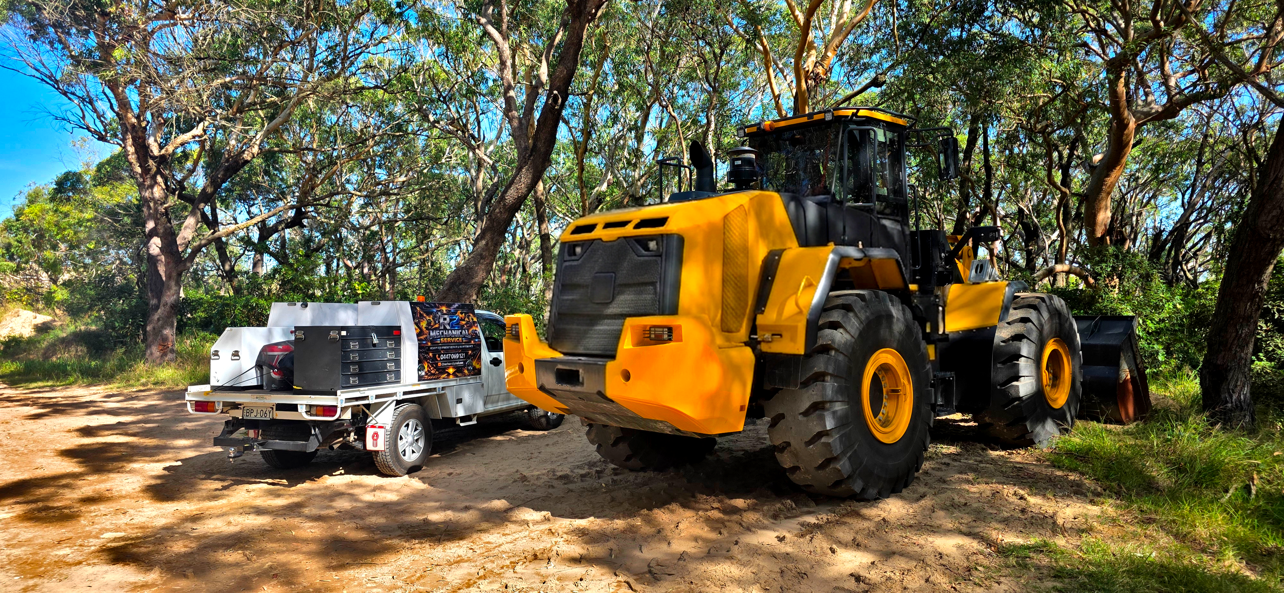 A yellow front loader parked on a dirt path, attached to a white utility trailer with tools and equipment, in a wooded area with tall trees and blue sky.
