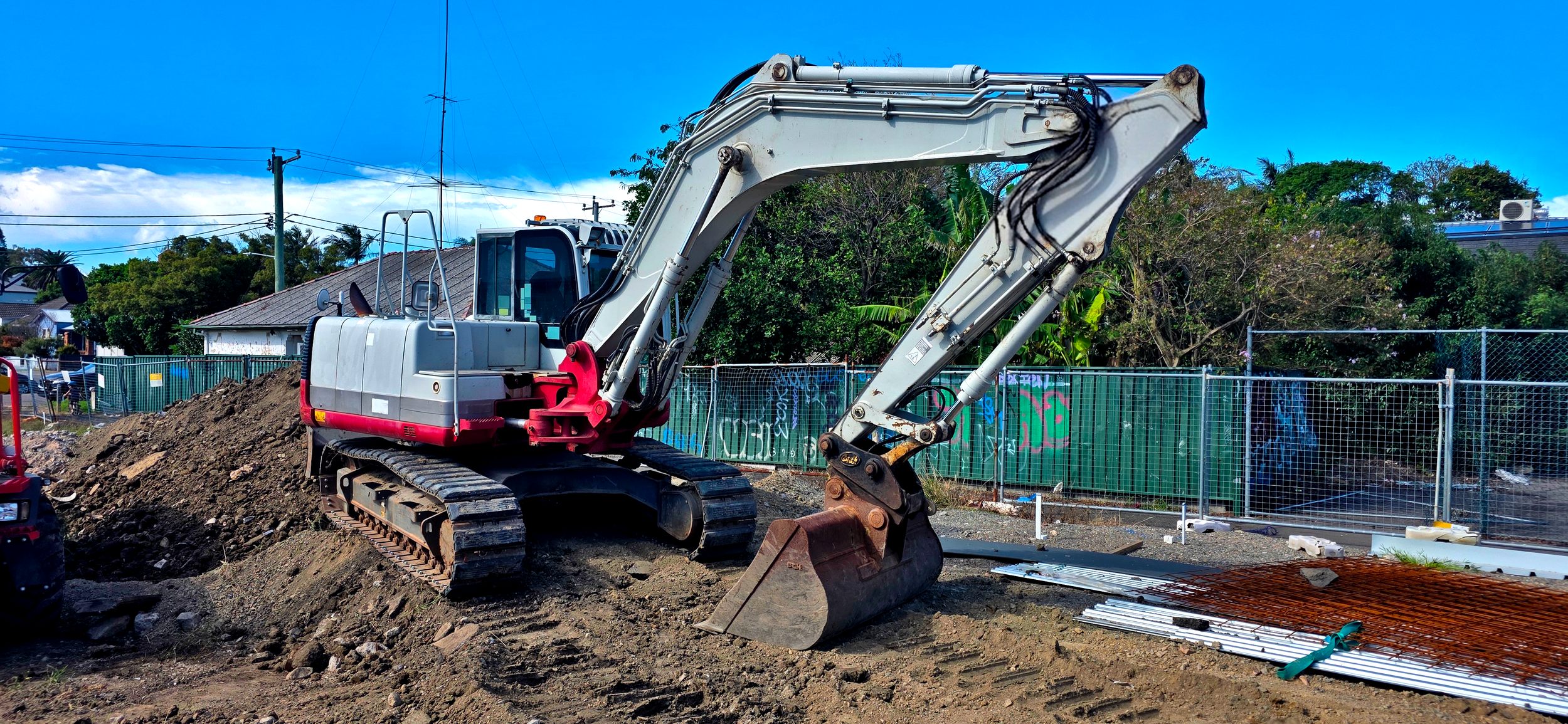 Construction site with a white and red excavator digging into dirt, surrounded by a fence with trees and houses in the background.