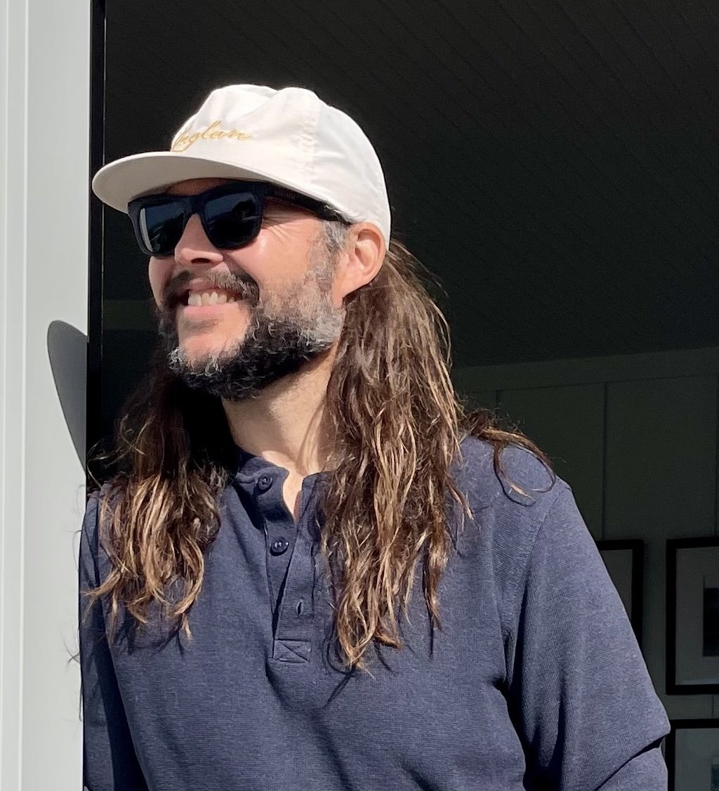 Smiling man with long hair and beard wearing a white cap, sunglasses, and a dark blue shirt, standing outside in bright sunlight.