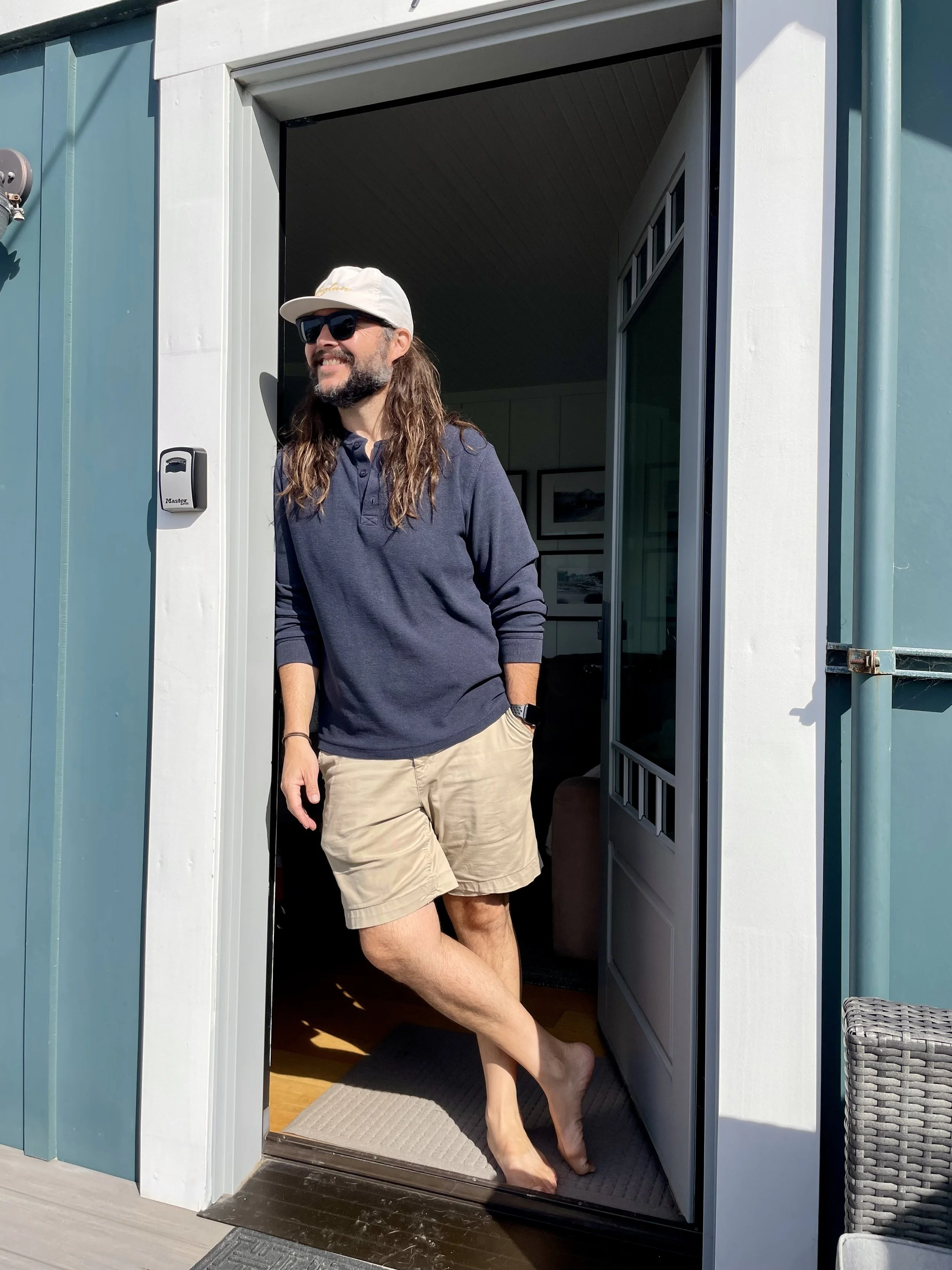 A man with long hair, beard, wearing sunglasses, a white cap, a navy polo shirt, and khaki shorts standing barefoot at the entrance of a house. He is smiling and looking outside, with one hand in his pocket and one foot slightly raised.
