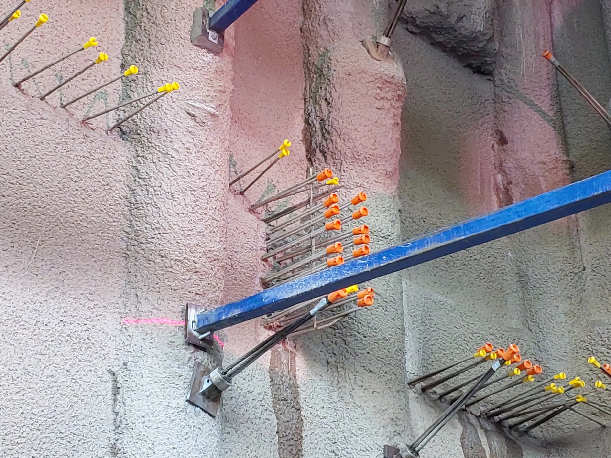 Construction site with steel rebar anchored in concrete, some with yellow and orange caps, and wooden supports, on a rough concrete surface.